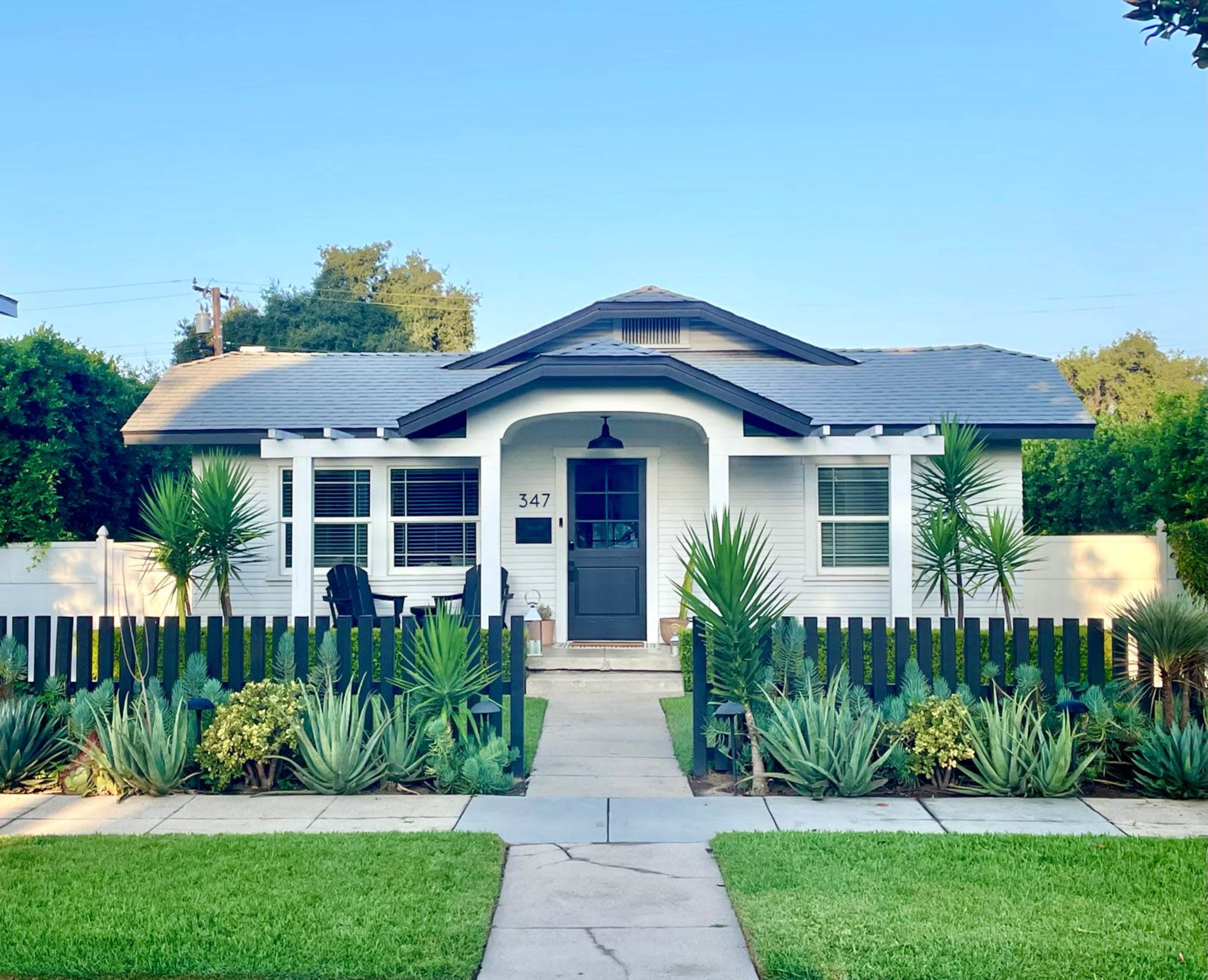 The image shows a white house with a gray roof and a black front door, surrounded by green foliage and a low black fence.