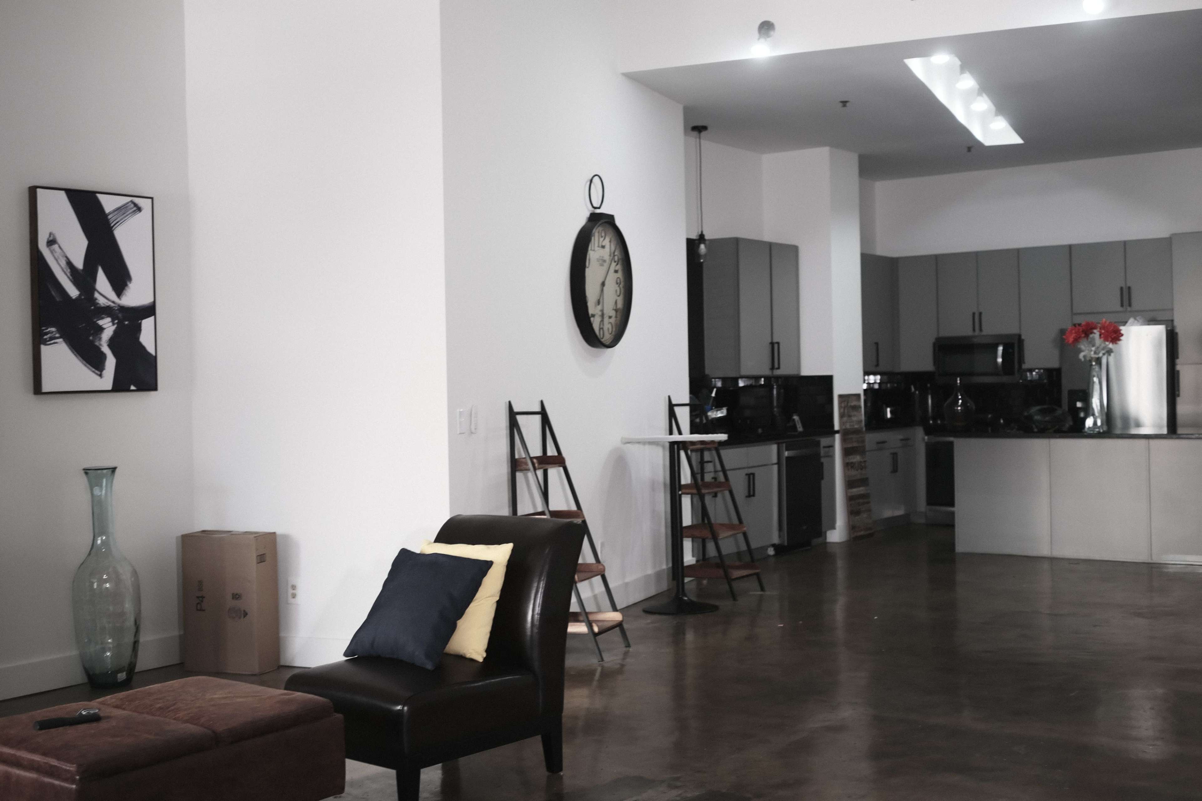 A modern open-concept living area featuring a dark leather chair, a coffee table, and a kitchen with gray cabinets and stainless steel appliances.