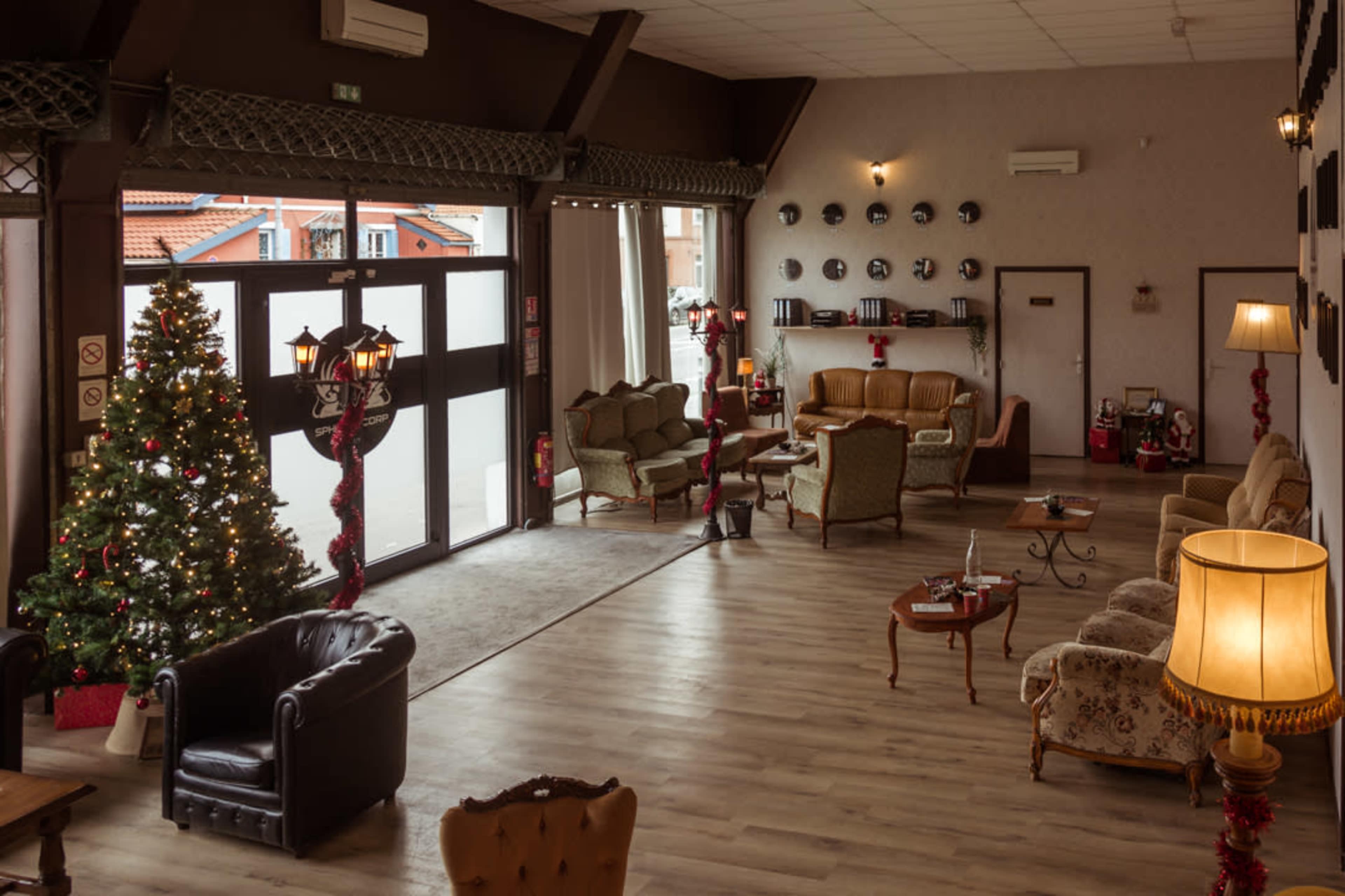 The image depicts a spacious lounge area decorated for the holidays, featuring a Christmas tree, various seating arrangements, and large windows allowing natural light to enter.