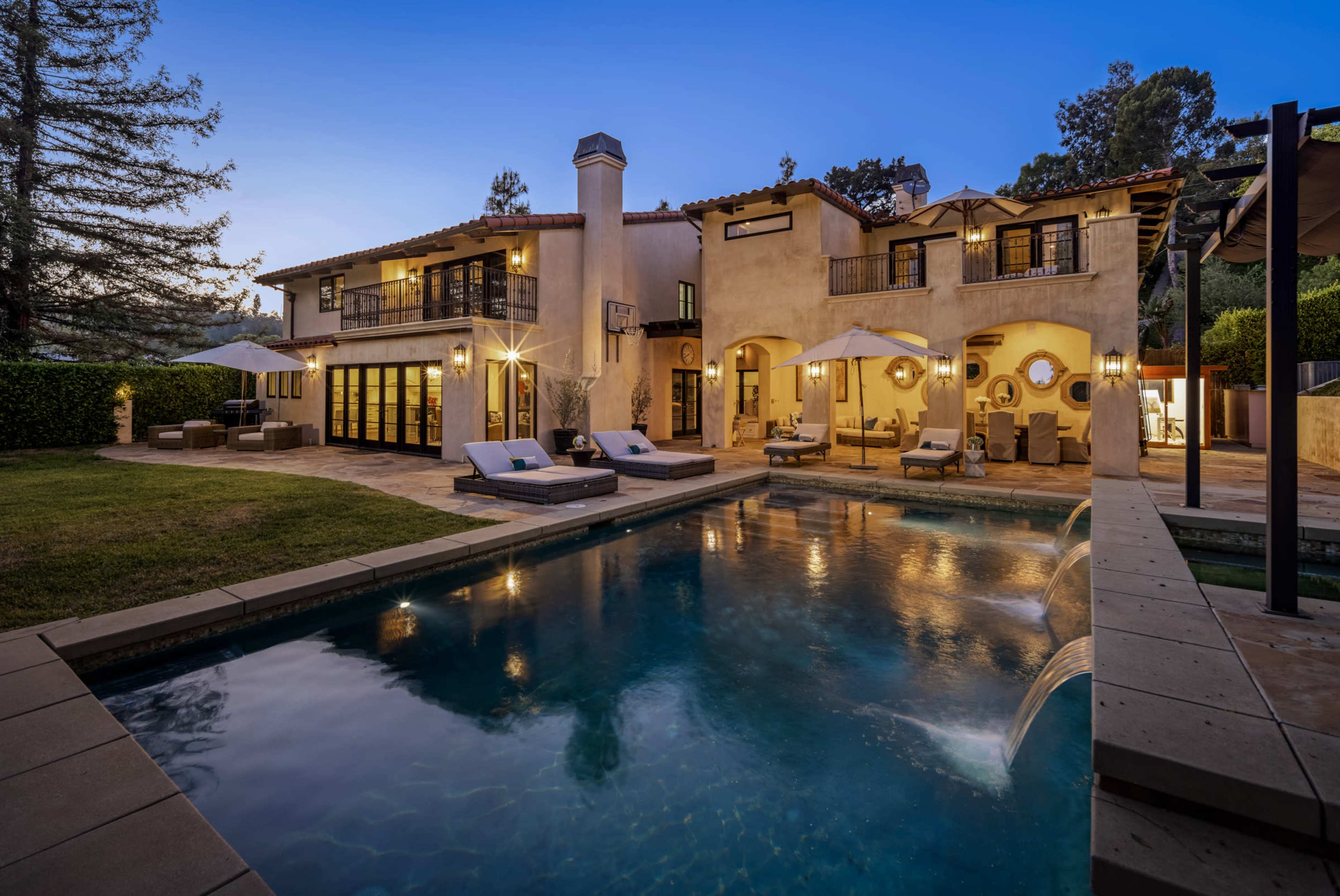 A two-story villa with a pool and outdoor seating areas, surrounded by greenery and illuminated in the evening light.