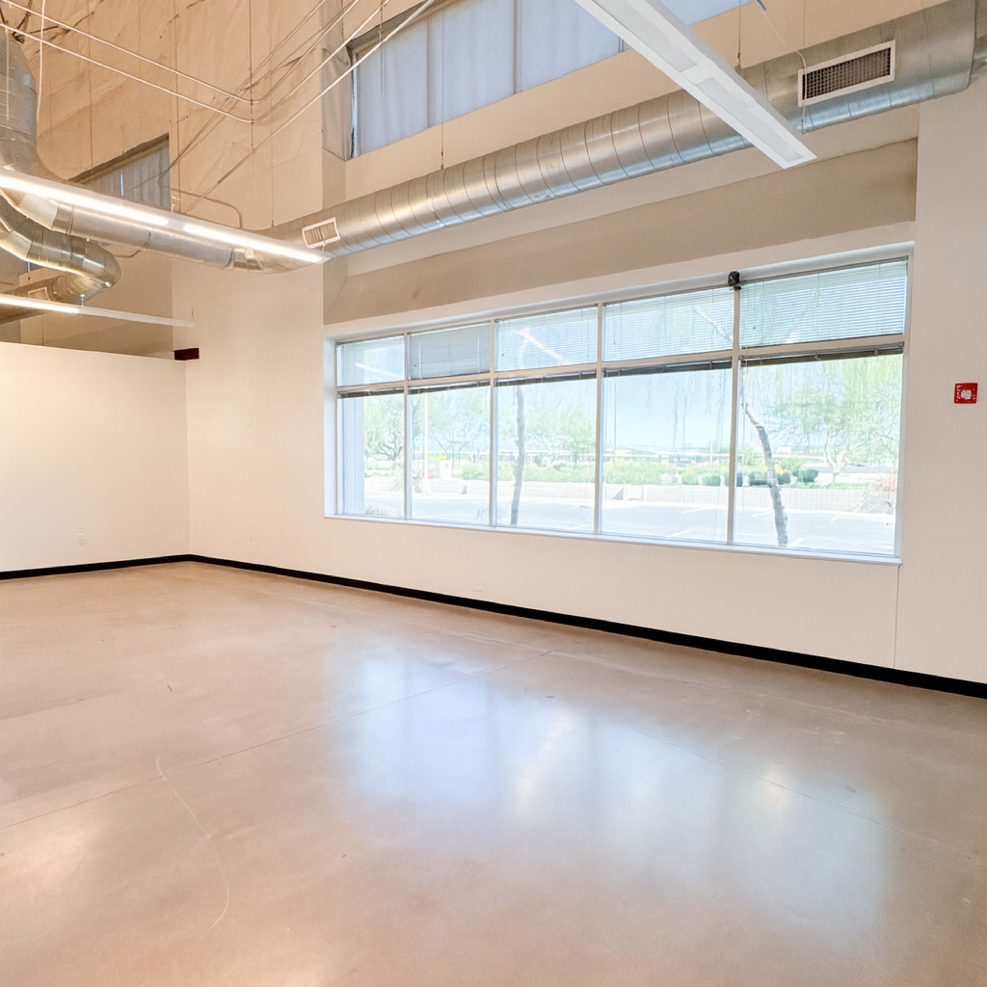 The image shows an empty room with large windows and industrial-style ductwork on the ceiling.