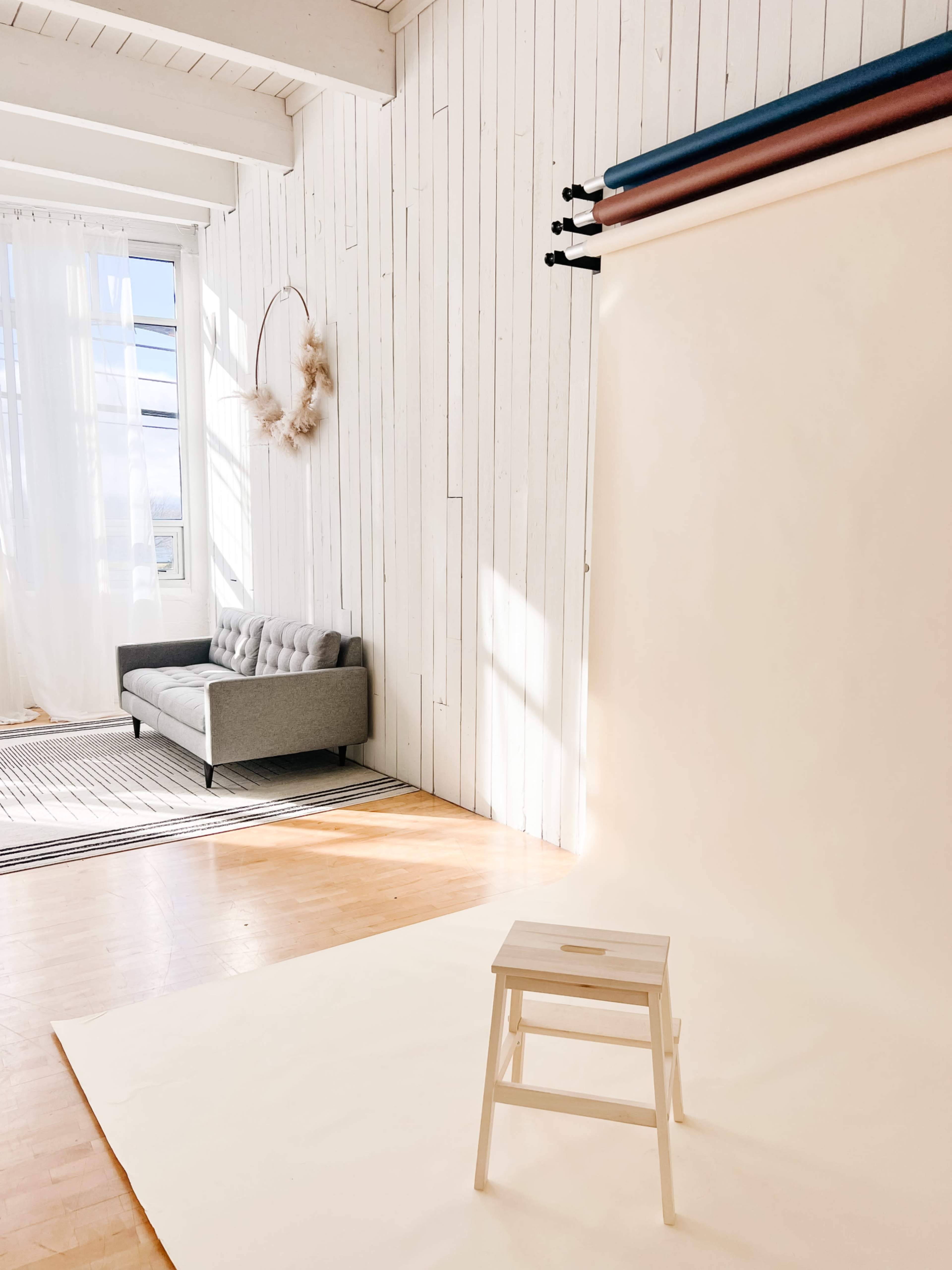 The scene features a bright, minimalist studio with a white wooden wall, a simple stool on a light backdrop, and a gray couch beside large windows allowing sunlight to fill the space.