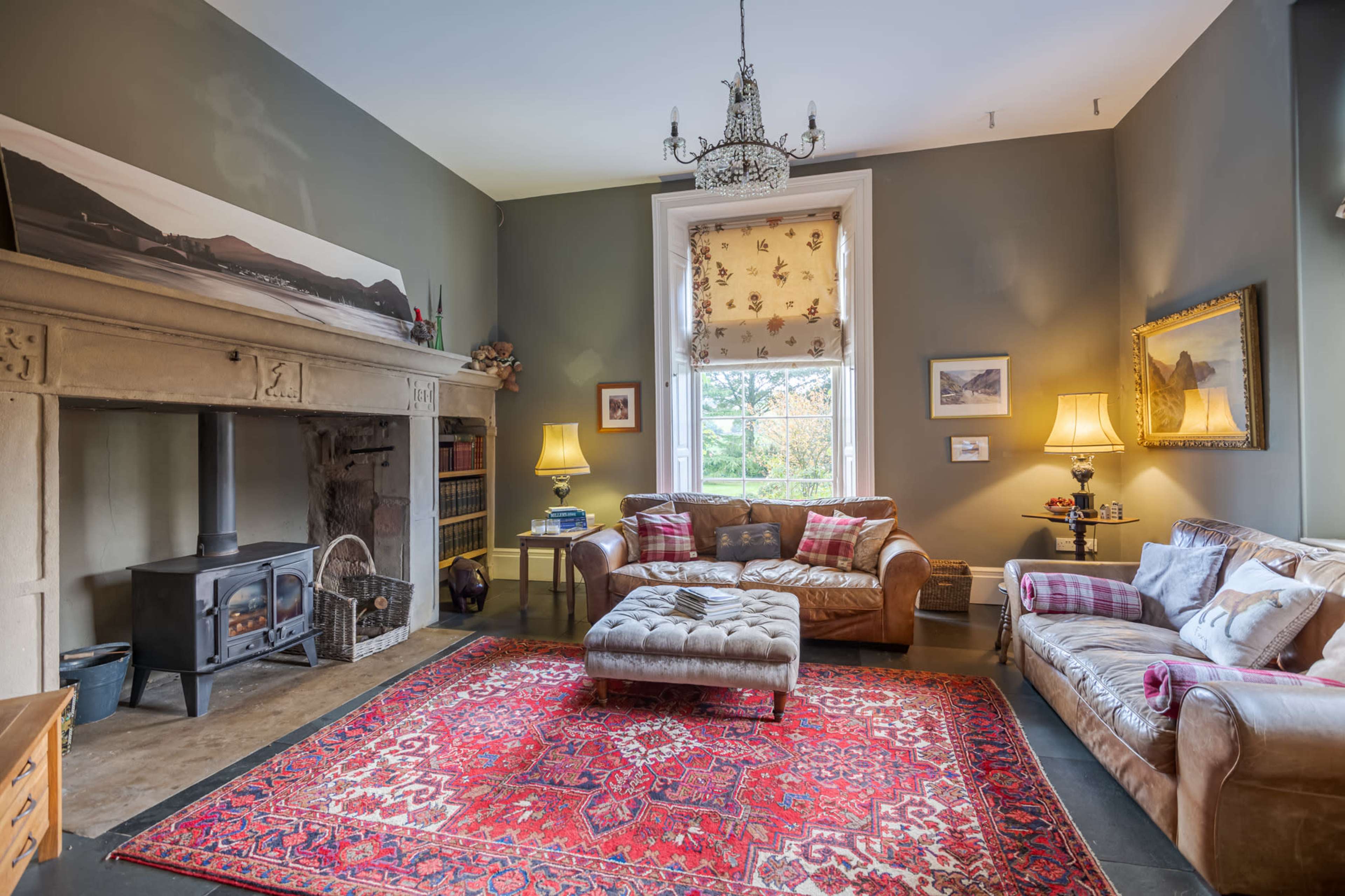 The image shows a cozy living room featuring a stone fireplace, a plush couch with decorative cushions, and a vibrant red area rug under a chandelier.