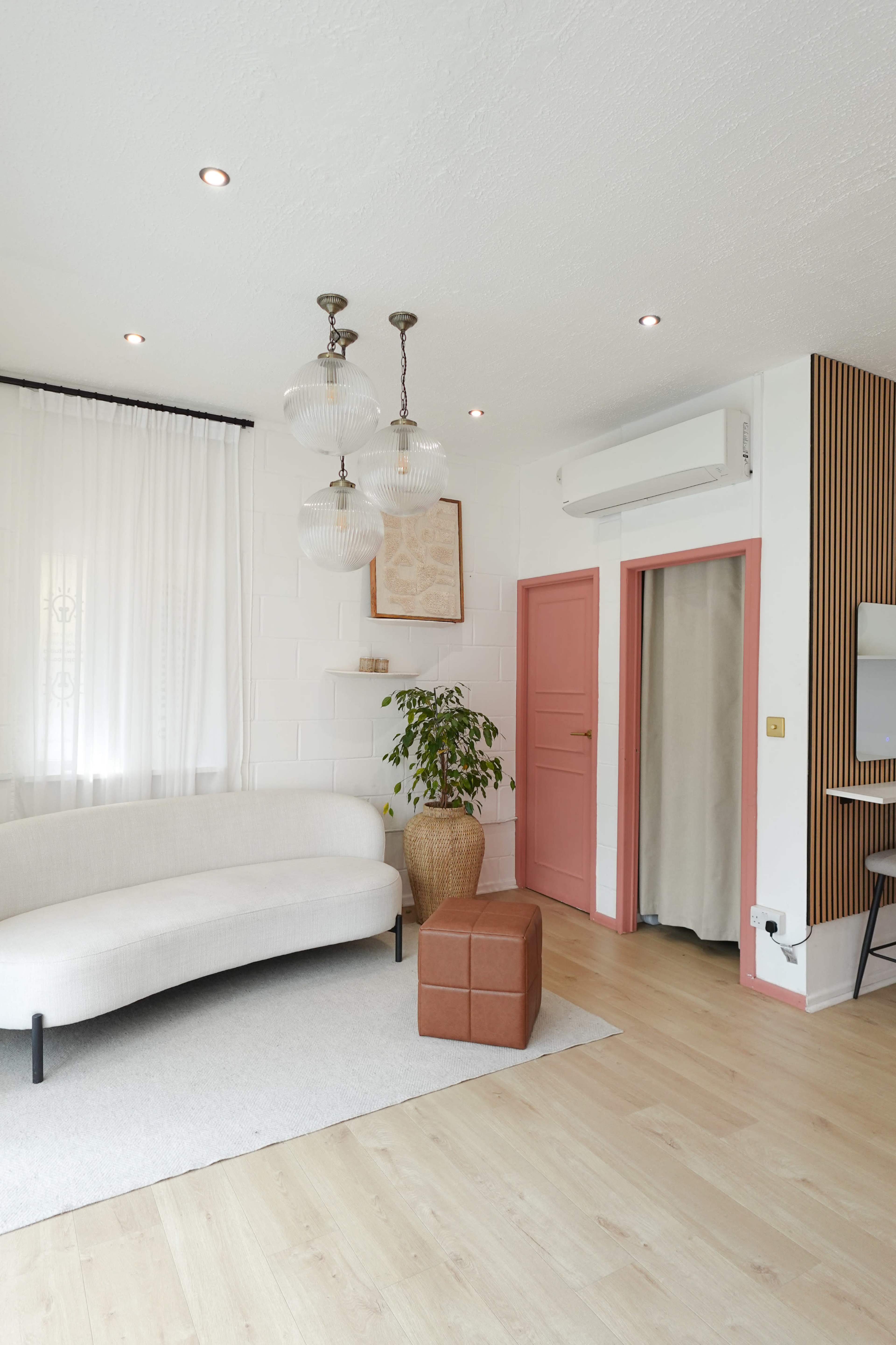 A modern living room features a white sofa, a brown ottoman, a leafy plant, and a pink door against a light-colored backdrop.