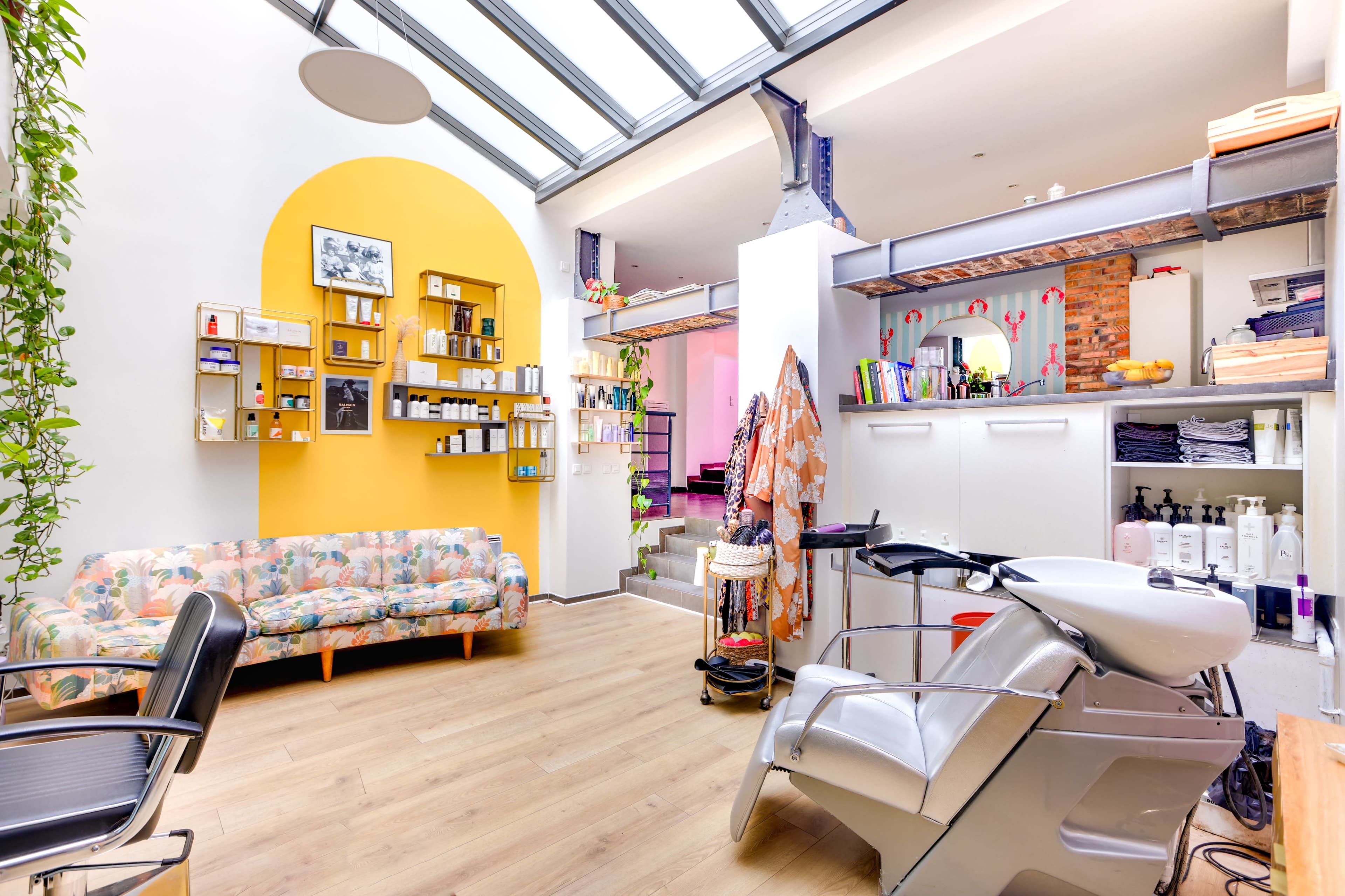 The image shows a bright and modern salon interior featuring a floral-patterned couch, a styling chair, and shelves displaying various hair and beauty products.