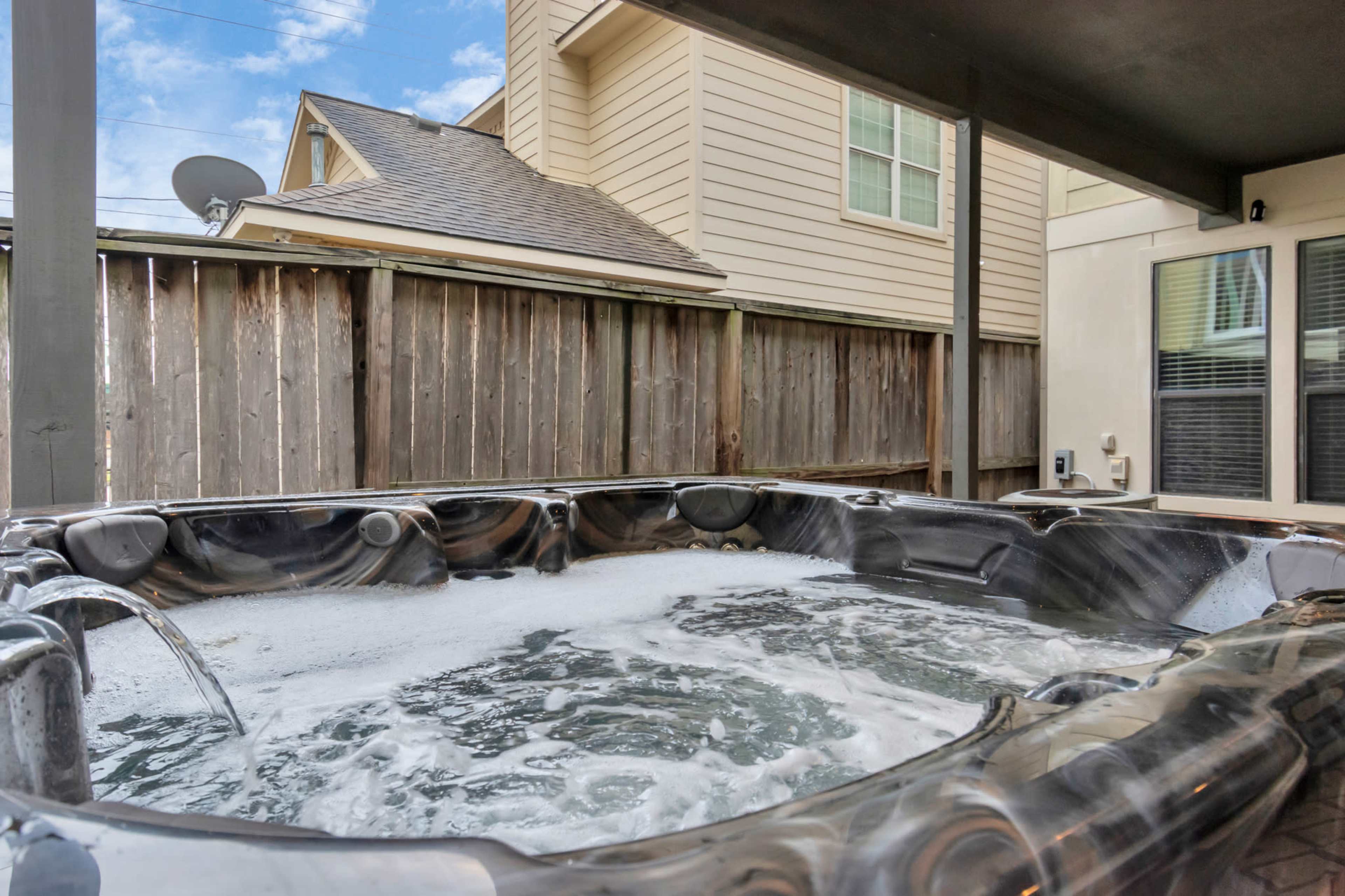 The image shows a hot tub filled with bubbling water, located on a patio next to a wooden fence and residential buildings.