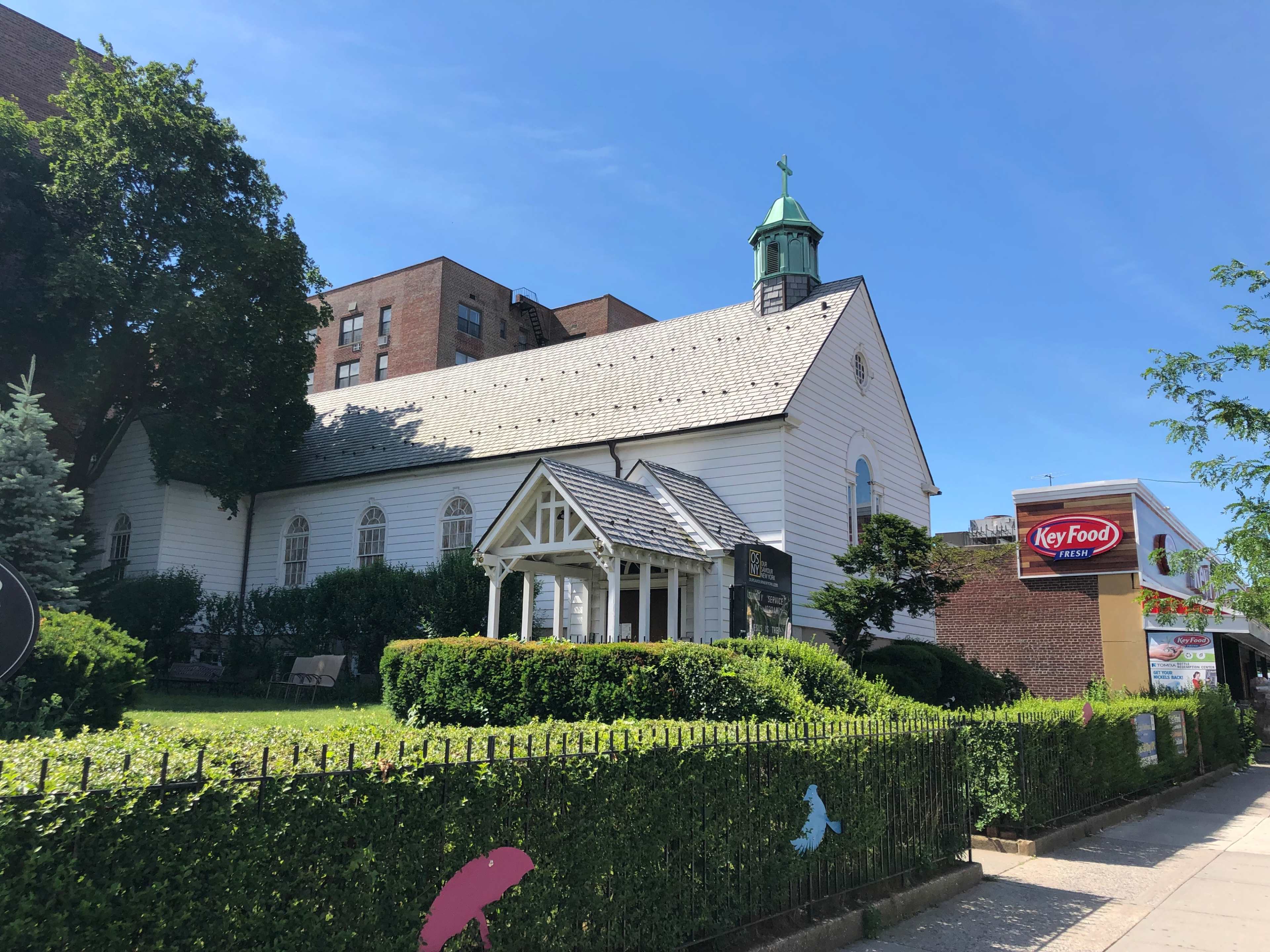 A white church with a green steeple is surrounded by greenery and is located next to a grocery store.