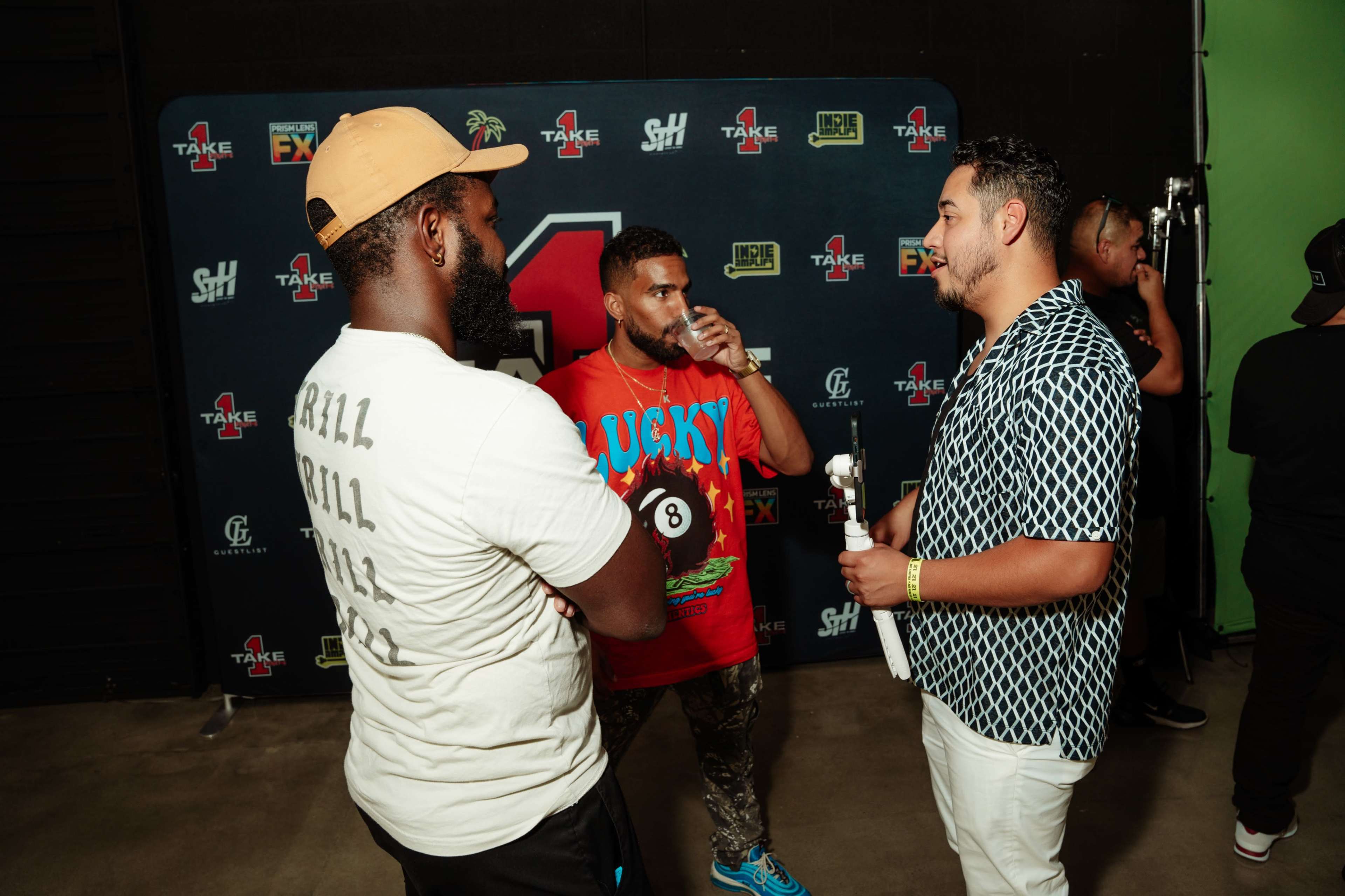 Three men are engaging in conversation, with one holding a drink, in front of a backdrop featuring the number one and various logos.