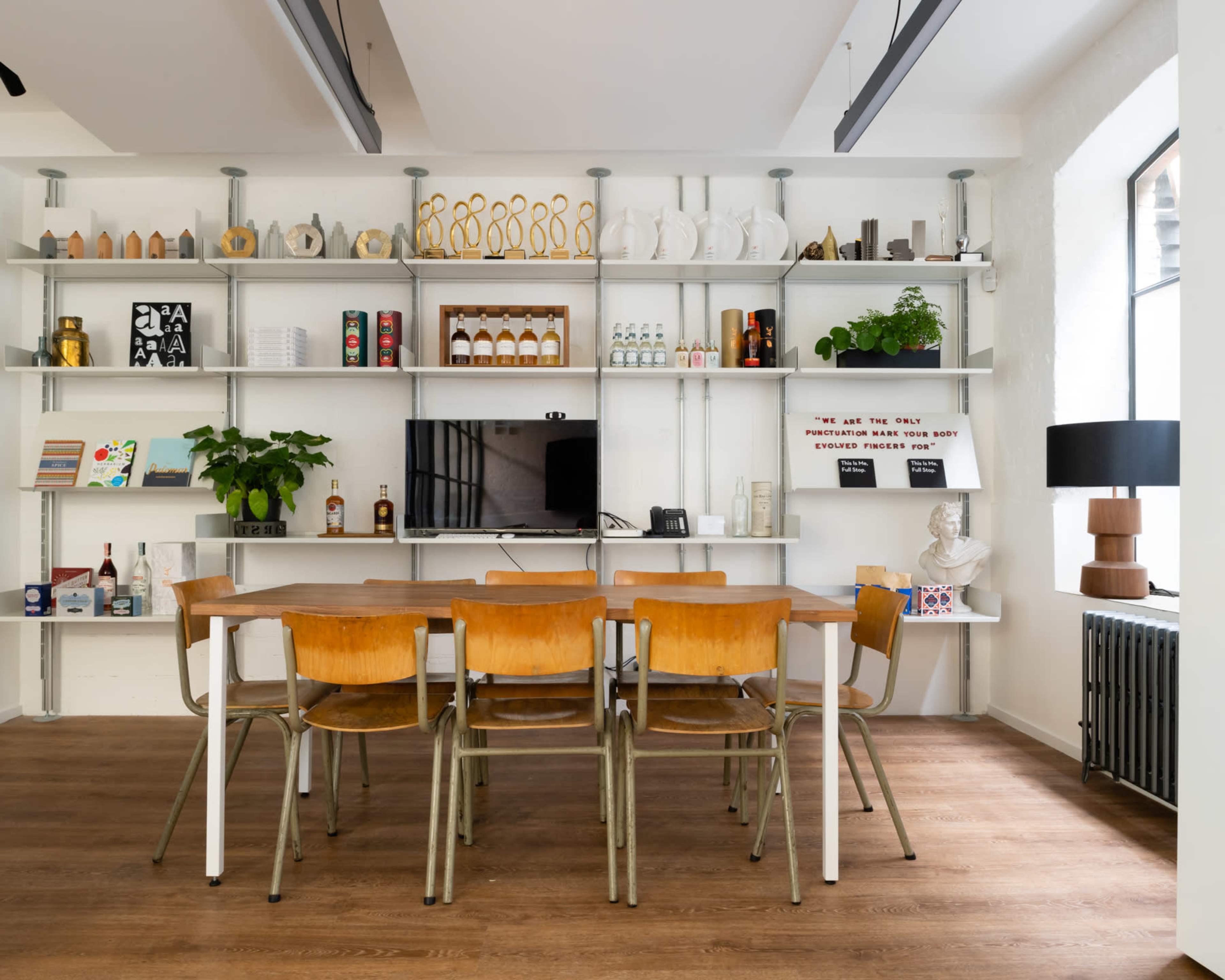 A modern workspace with a wooden table surrounded by chairs, and shelves displaying various decorative items and bottles.