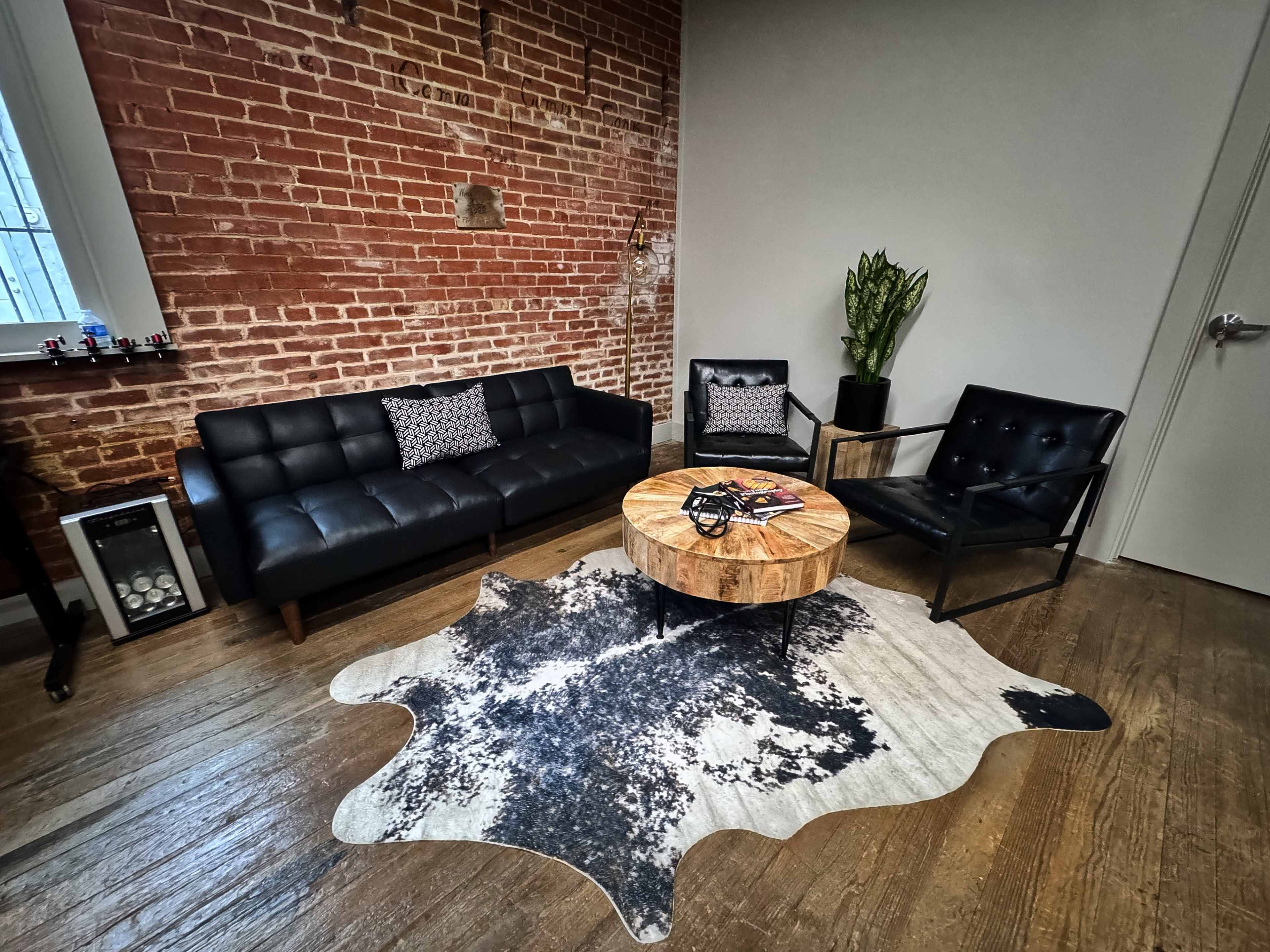 A modern living area with a black leather sofa, two black chairs, a round wooden coffee table, and a cowhide rug on wooden flooring, against a backdrop of exposed red brick walls.
