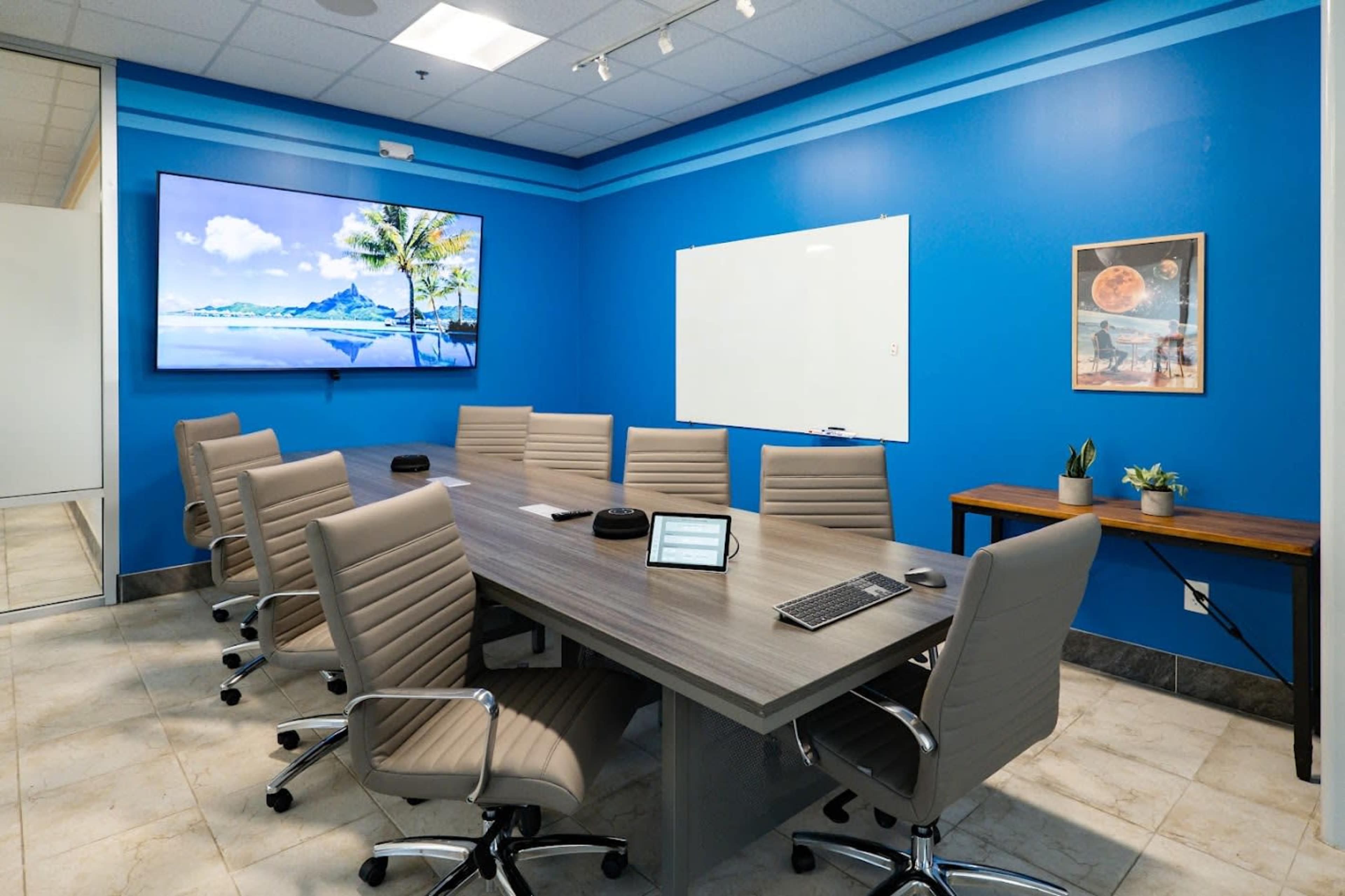 A modern conference room features a long table surrounded by ergonomic chairs, a large screen on the wall, and a whiteboard.