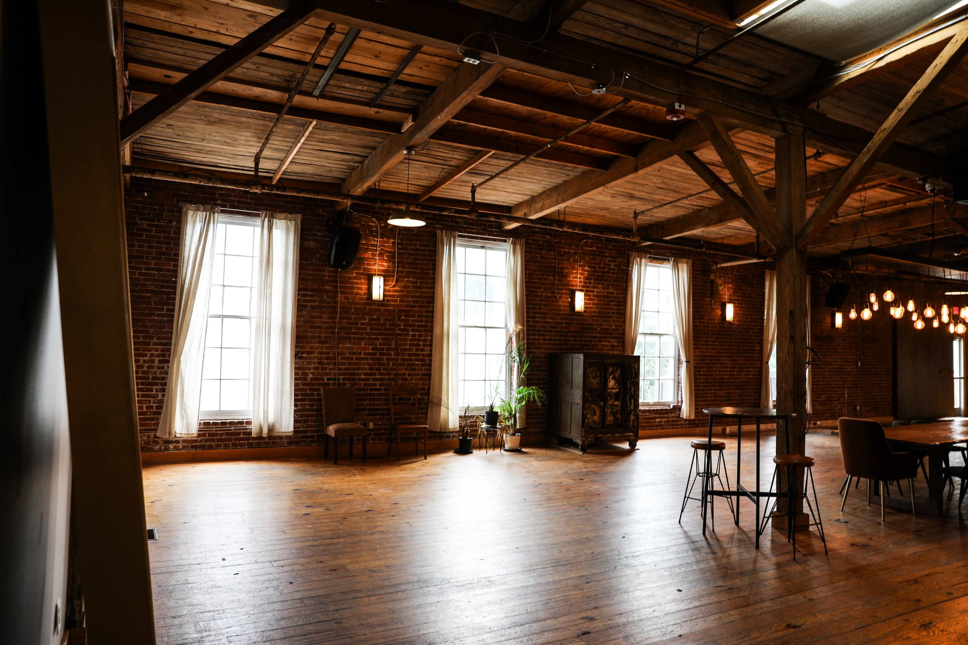A spacious room features exposed brick walls, large windows, wooden beams, and various seating arrangements.