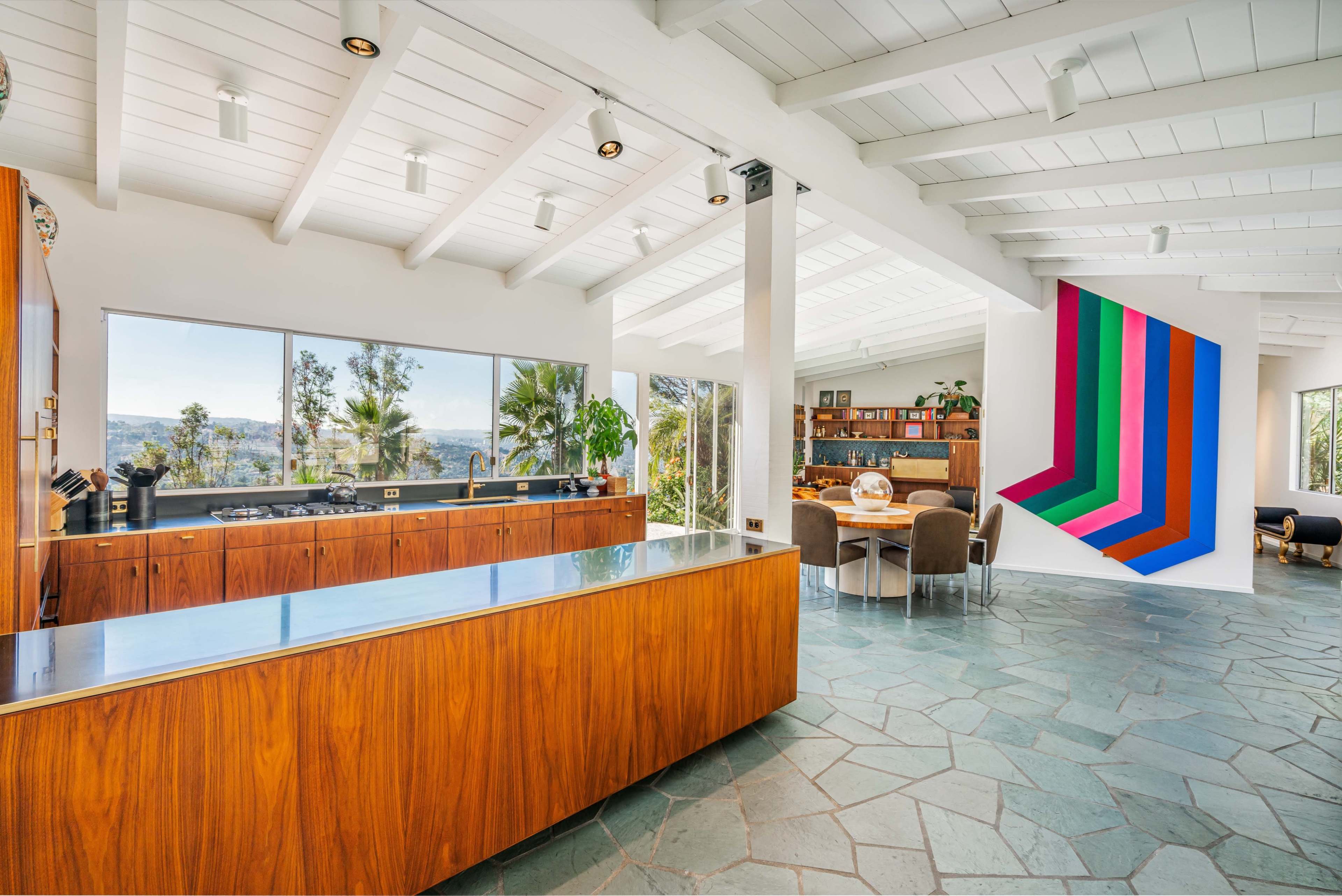 A modern kitchen with wooden cabinetry, a large island, and a dining area featuring a geometric artwork on the wall and large windows showing a view of greenery outside.