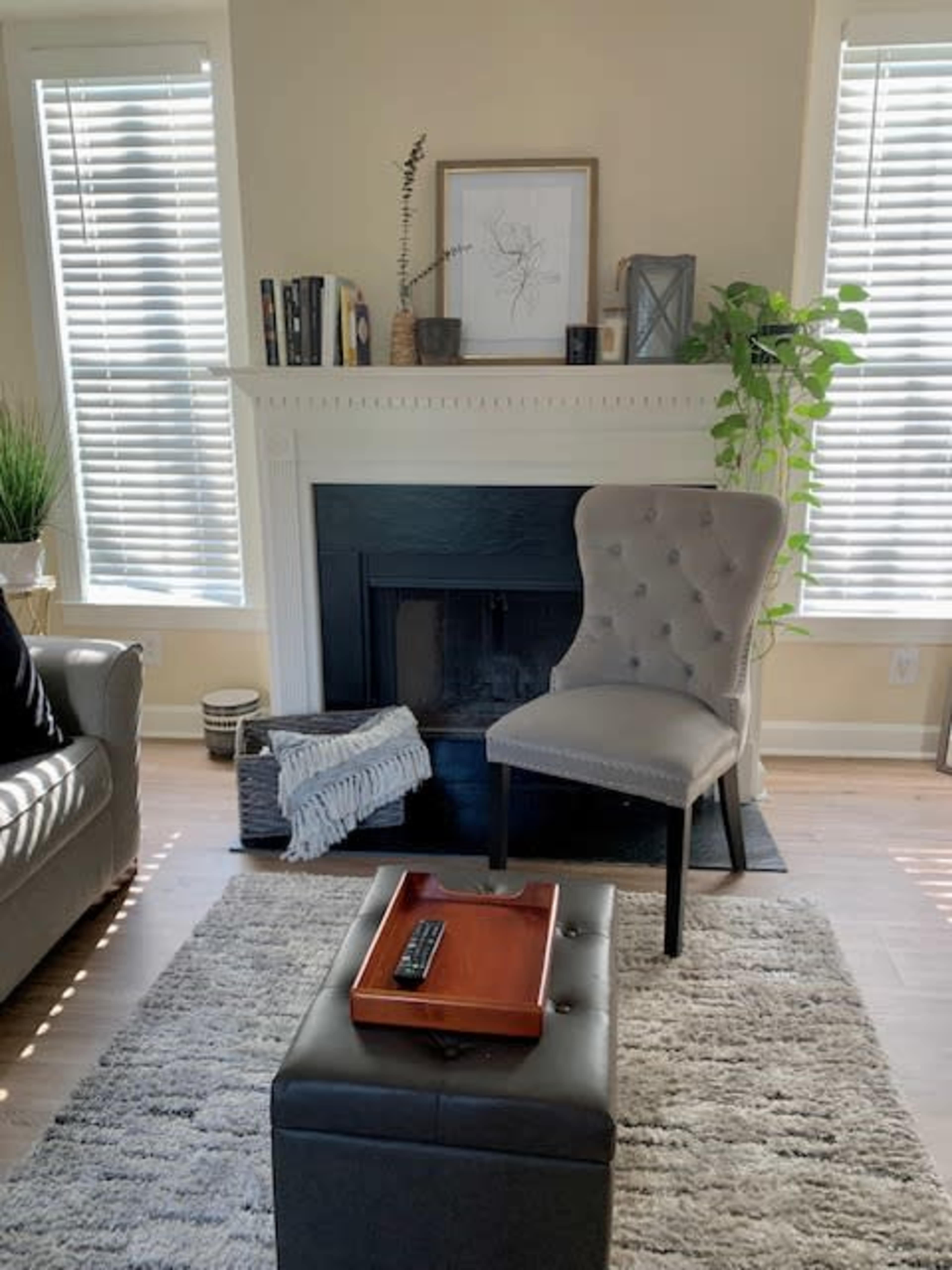 A living room with a tufted chair positioned near a fireplace, surrounded by large windows, a coffee table with a tray, and a decorative plant.