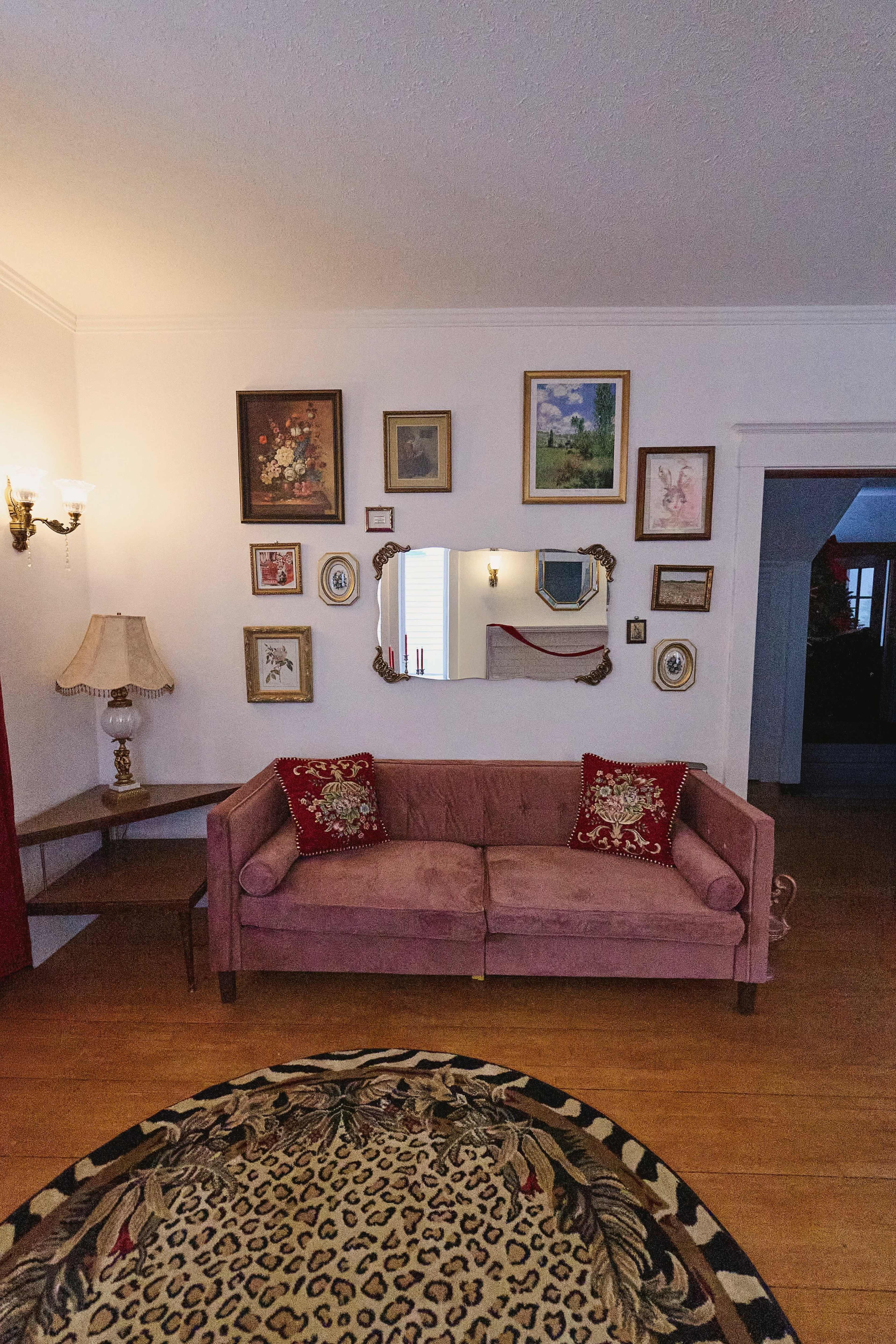 The image shows a cozy living room featuring a pink couch, various framed artworks on the walls, and a decorative round rug with a leopard print.
