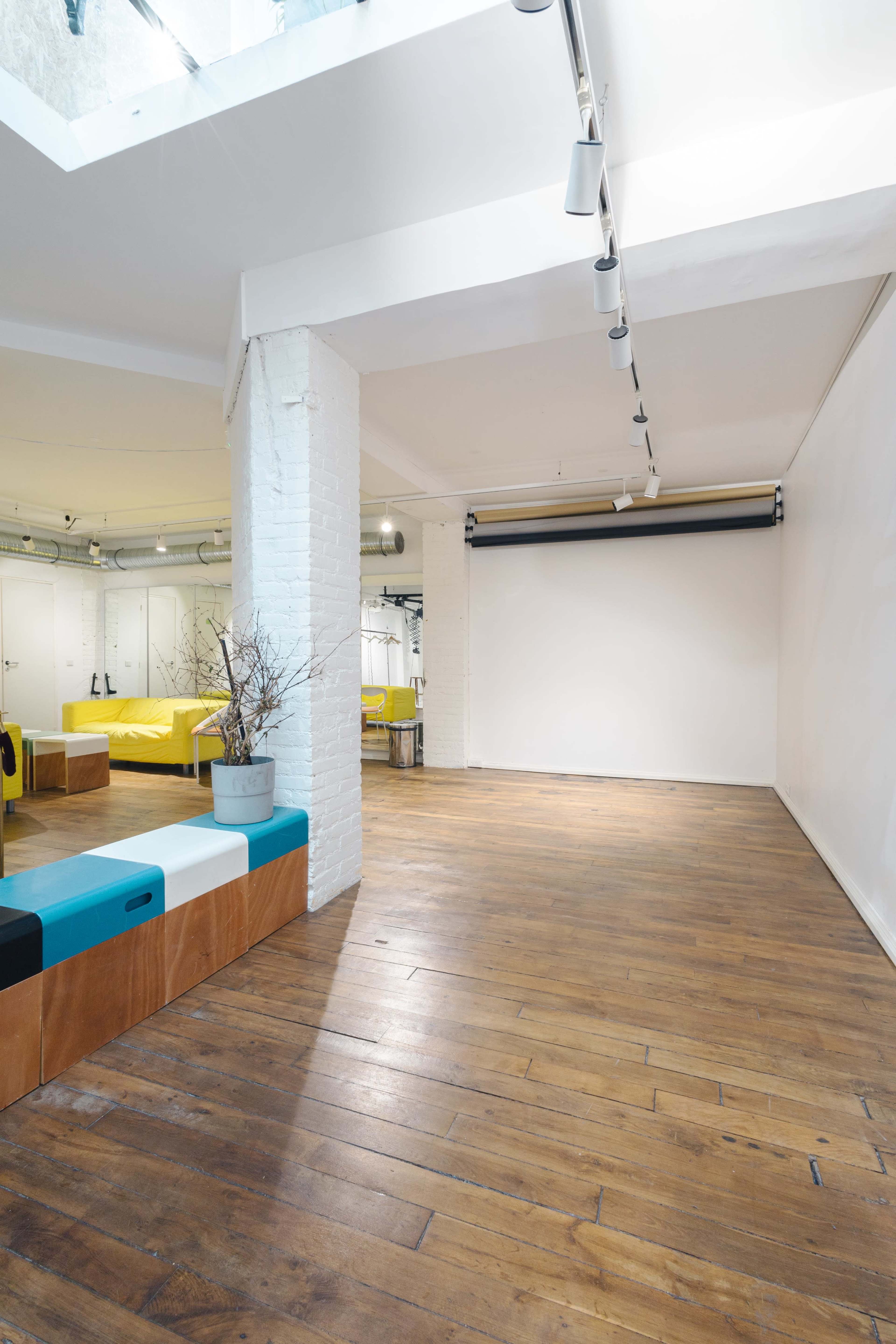 A spacious interior with wooden floors, white walls, and a few pieces of minimalist furniture, including a colorful bench and a large blank wall ideal for presentations.