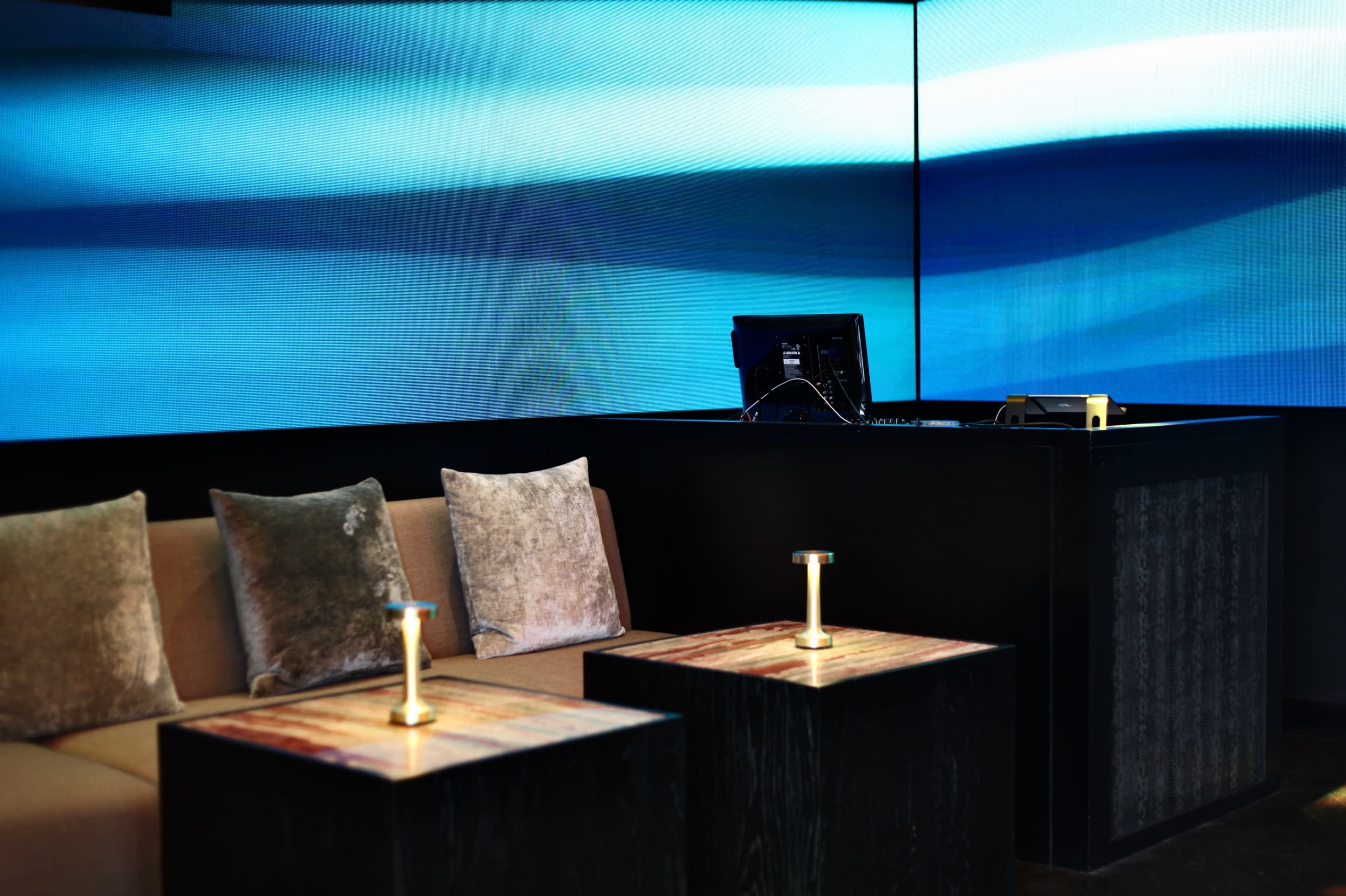 A modern lounge area with a sofa and two tables, accompanied by a computer setup against a backdrop of large blue screens.