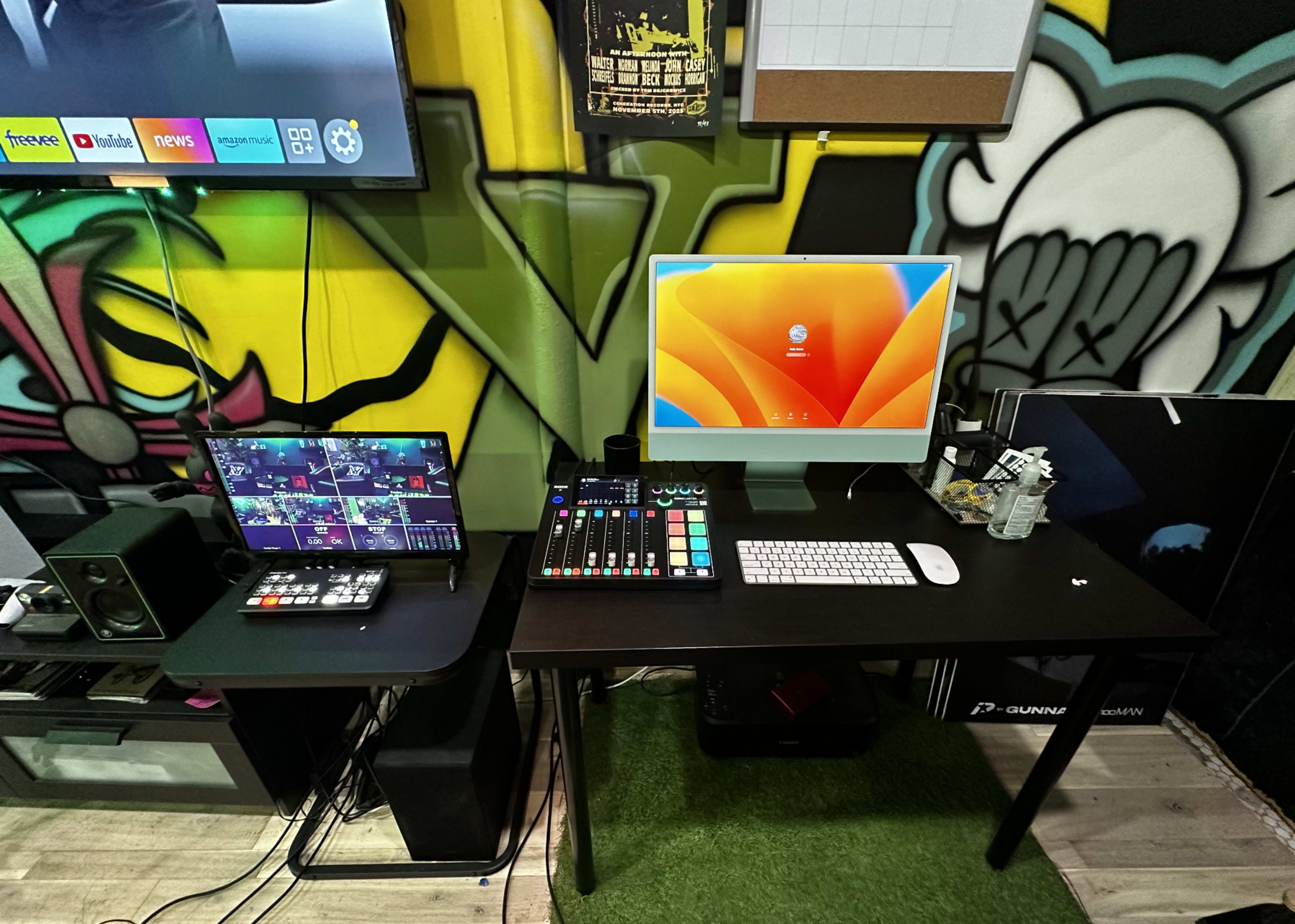 A black desk with a computer monitor, a laptop, a soundboard, a keyboard, and a water bottle against a colorful graffiti-style wall.