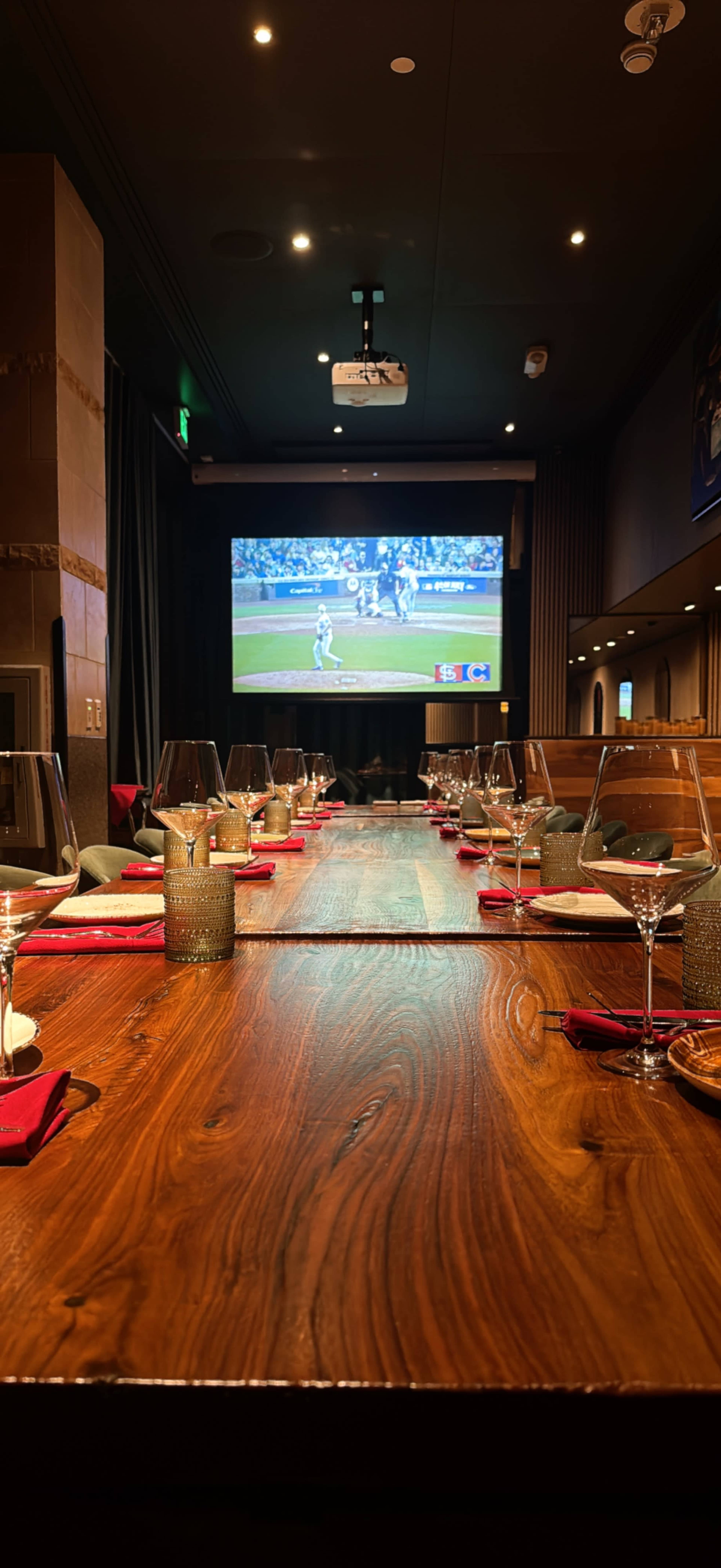 A long wooden dining table is set with glassware and red napkins, facing a large screen displaying a baseball game.