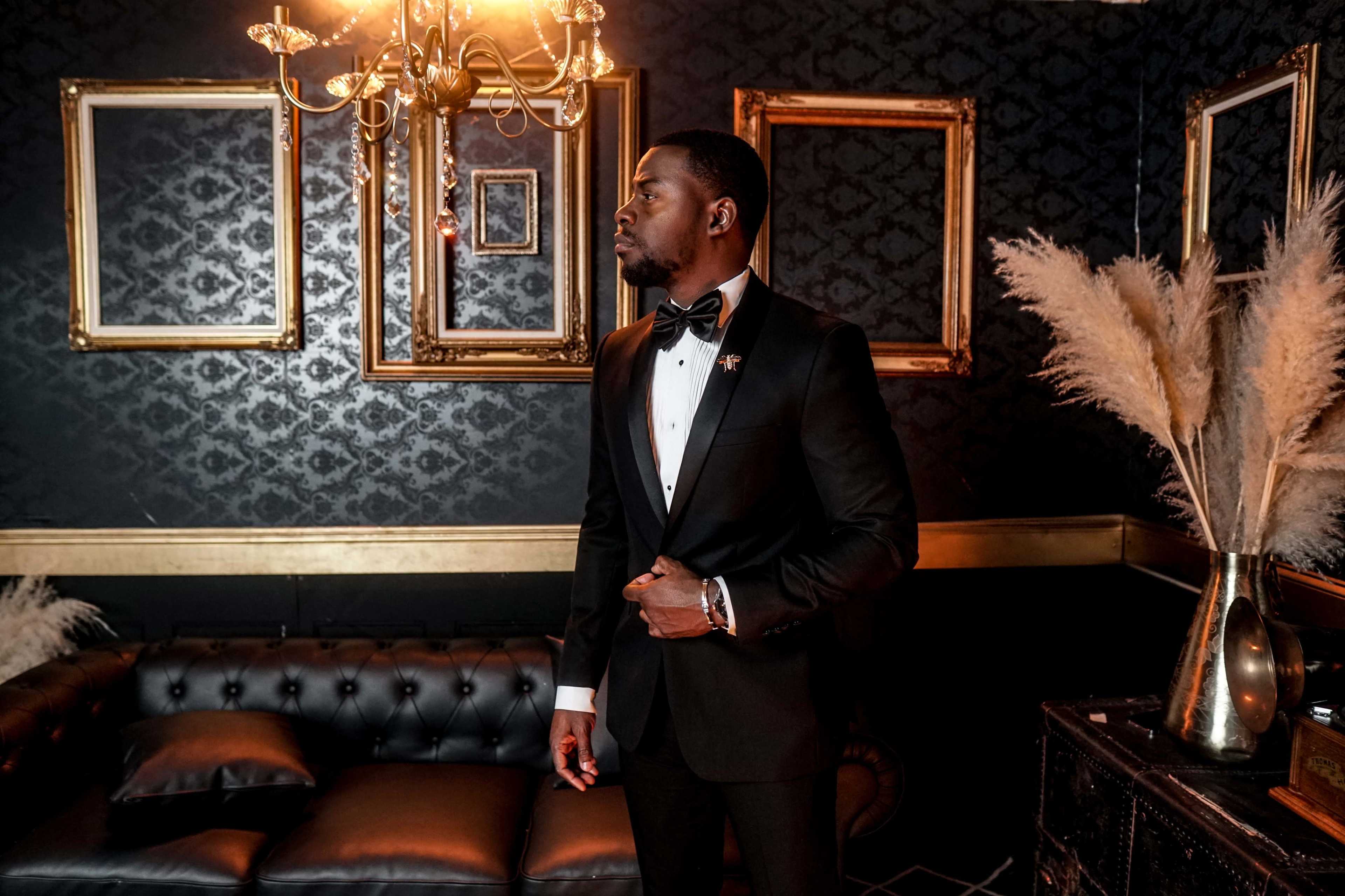 A man in a black tuxedo stands beside a leather couch in a dimly lit room with decorative gold frames and a chandelier.