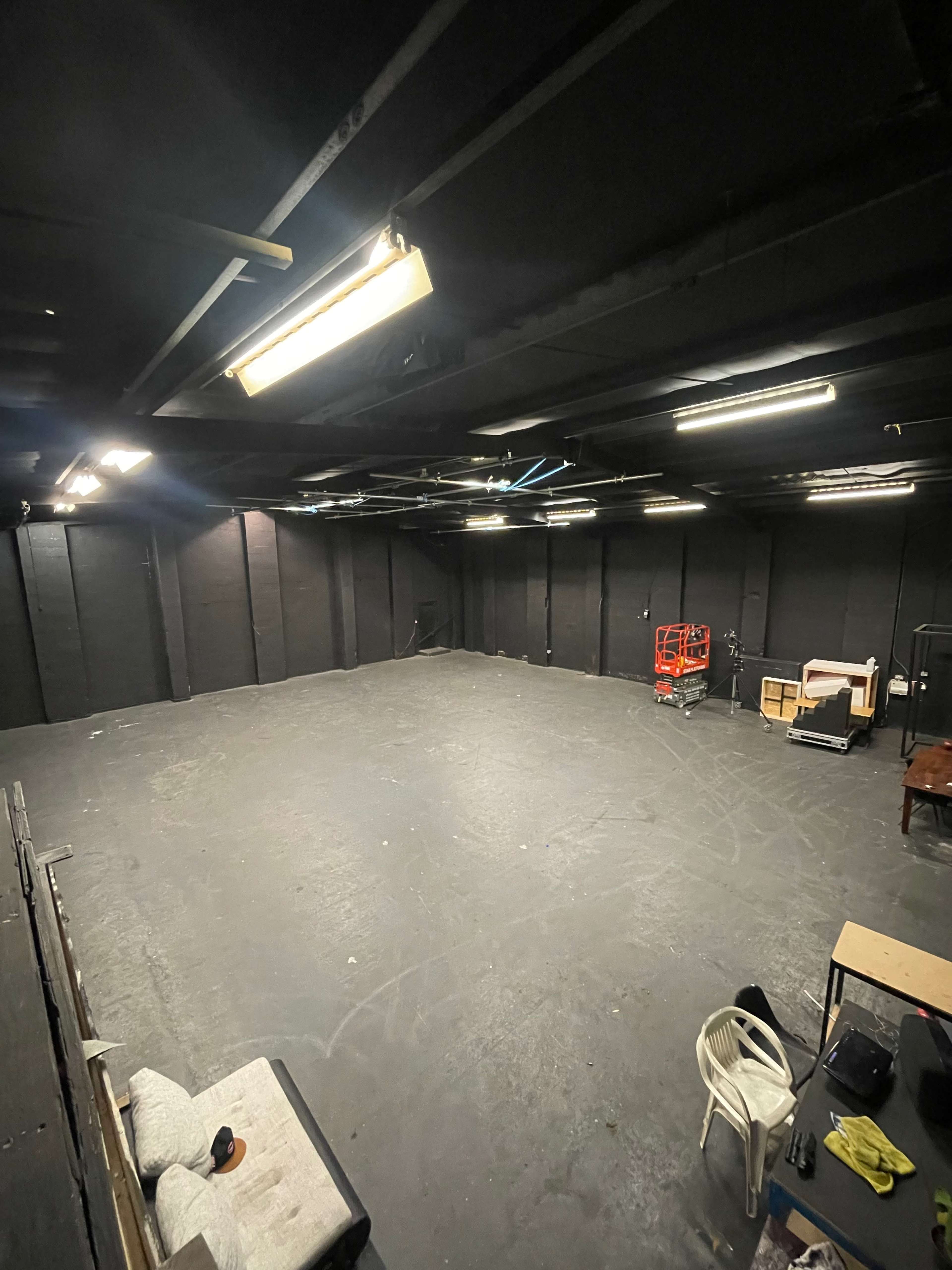 A large, empty room with black walls and a concrete floor, featuring minimal furniture and overhead lighting.