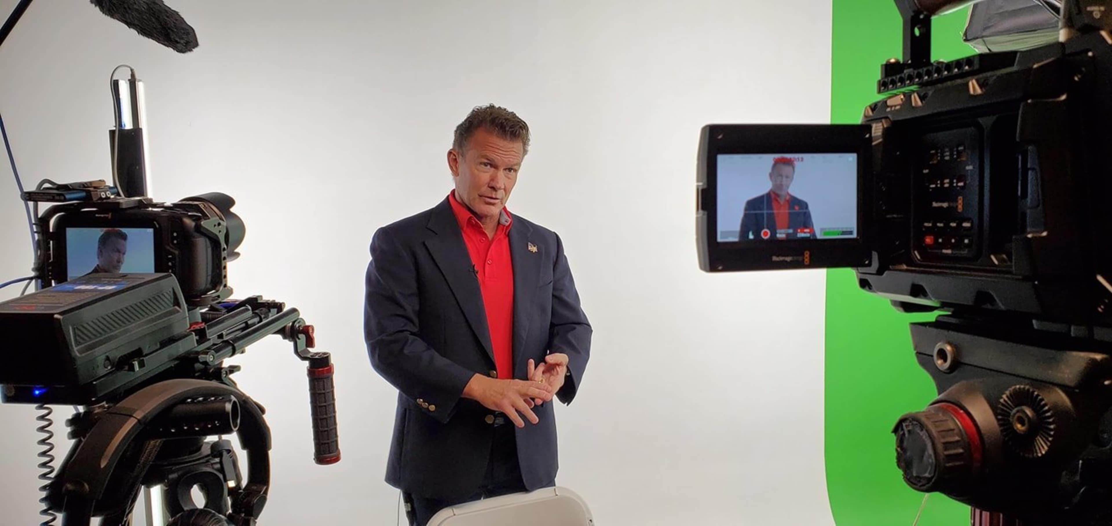 A presenter gestures with his hands in front of a camera setup against a green and white backdrop.