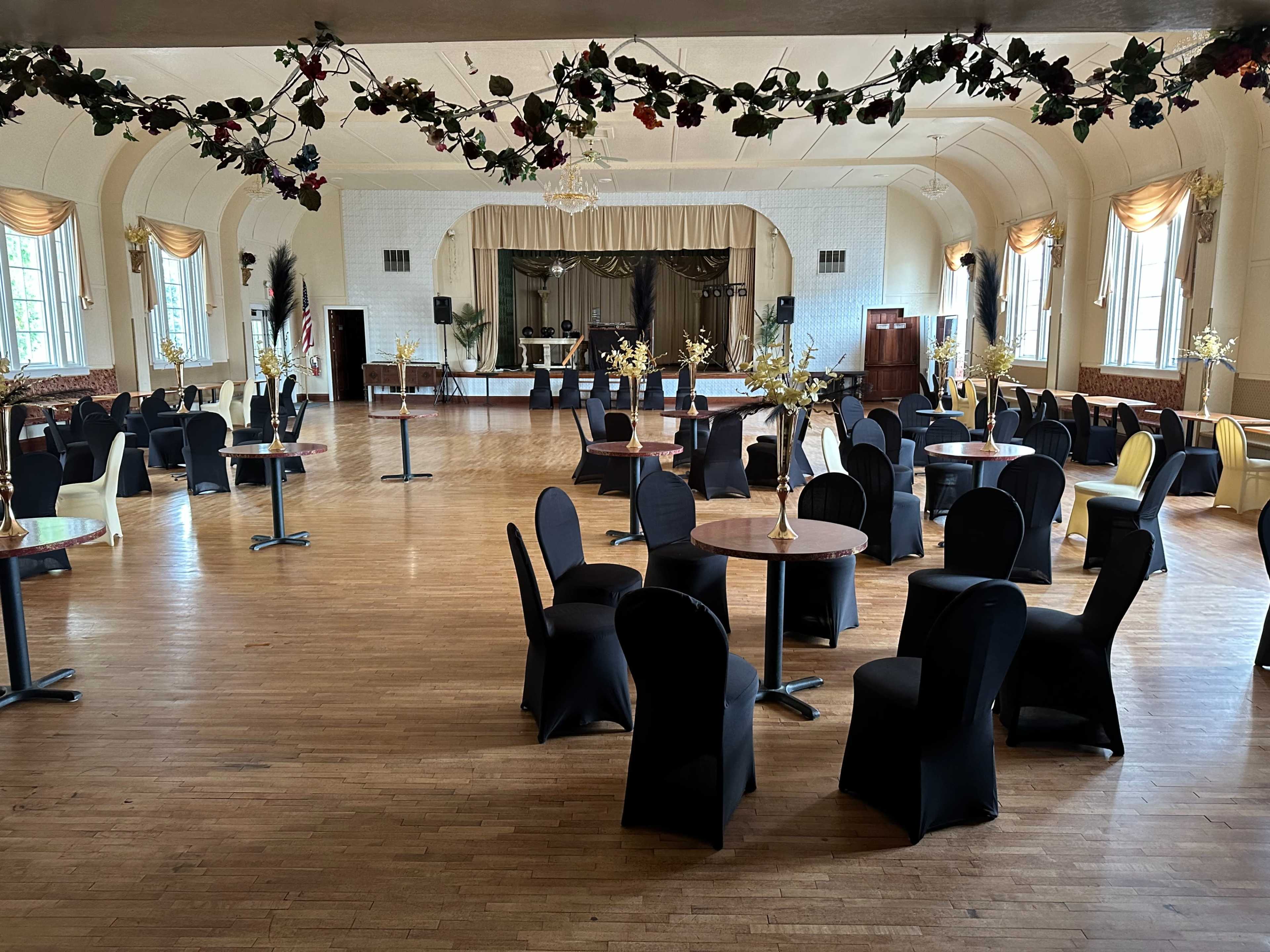 A large, empty banquet hall with neatly arranged tables and chairs, a stage in the background, and decorative elements on the ceiling.