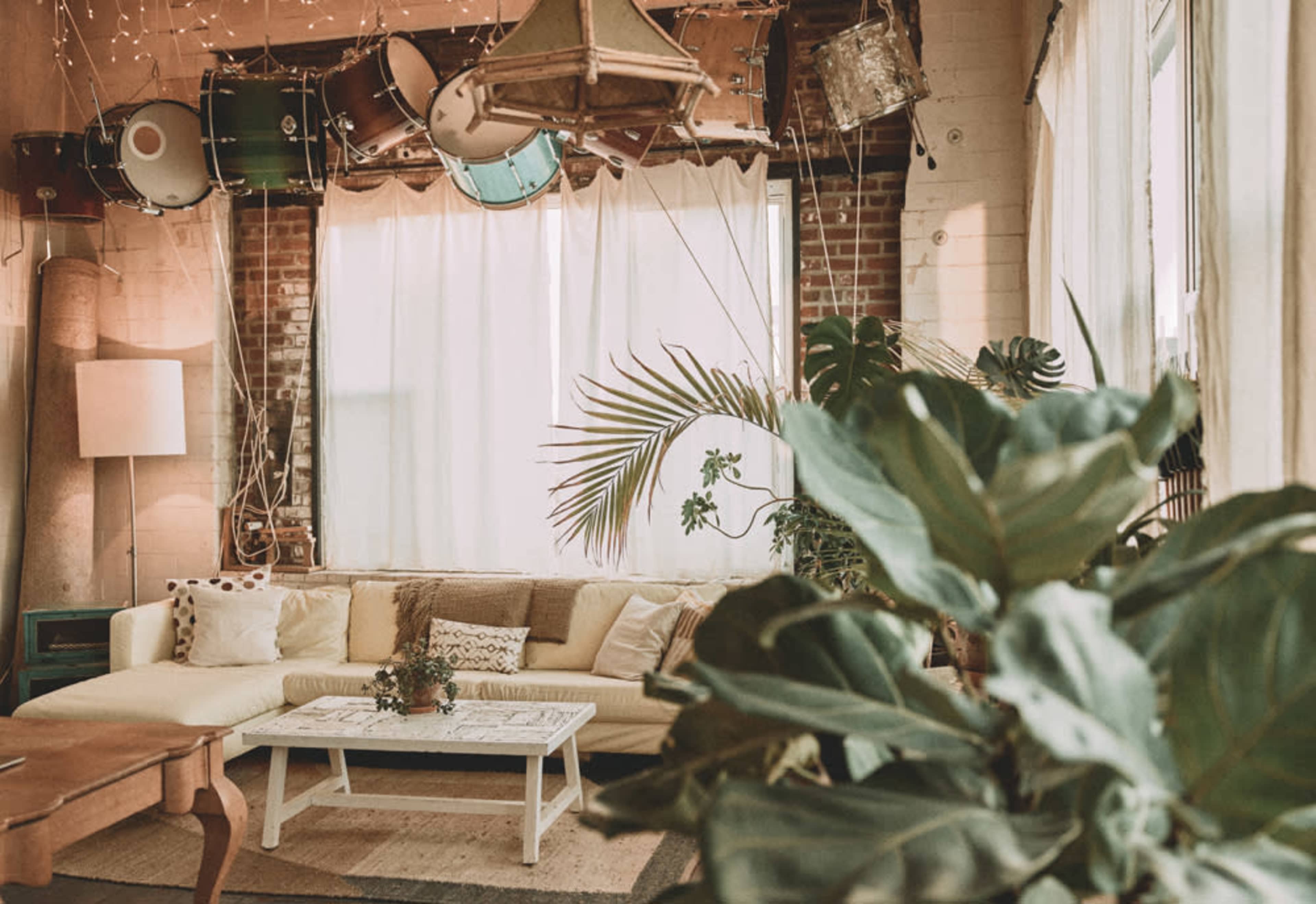 A cozy living room with a white sofa, a coffee table, and plants, featuring a backdrop of large windows and hanging drums.