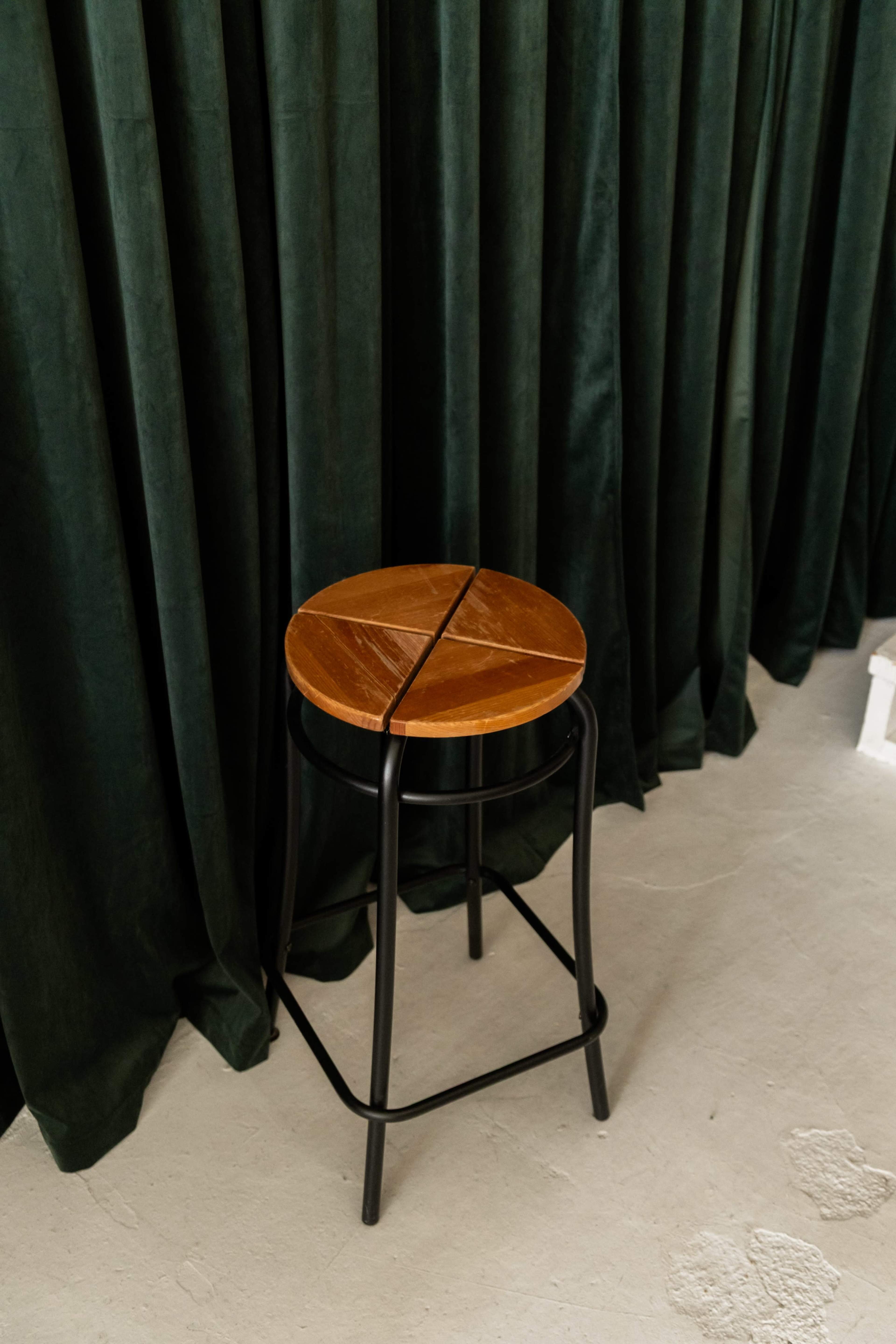 A wooden stool with a split circular seat stands in front of dark green curtains on a light-colored floor.
