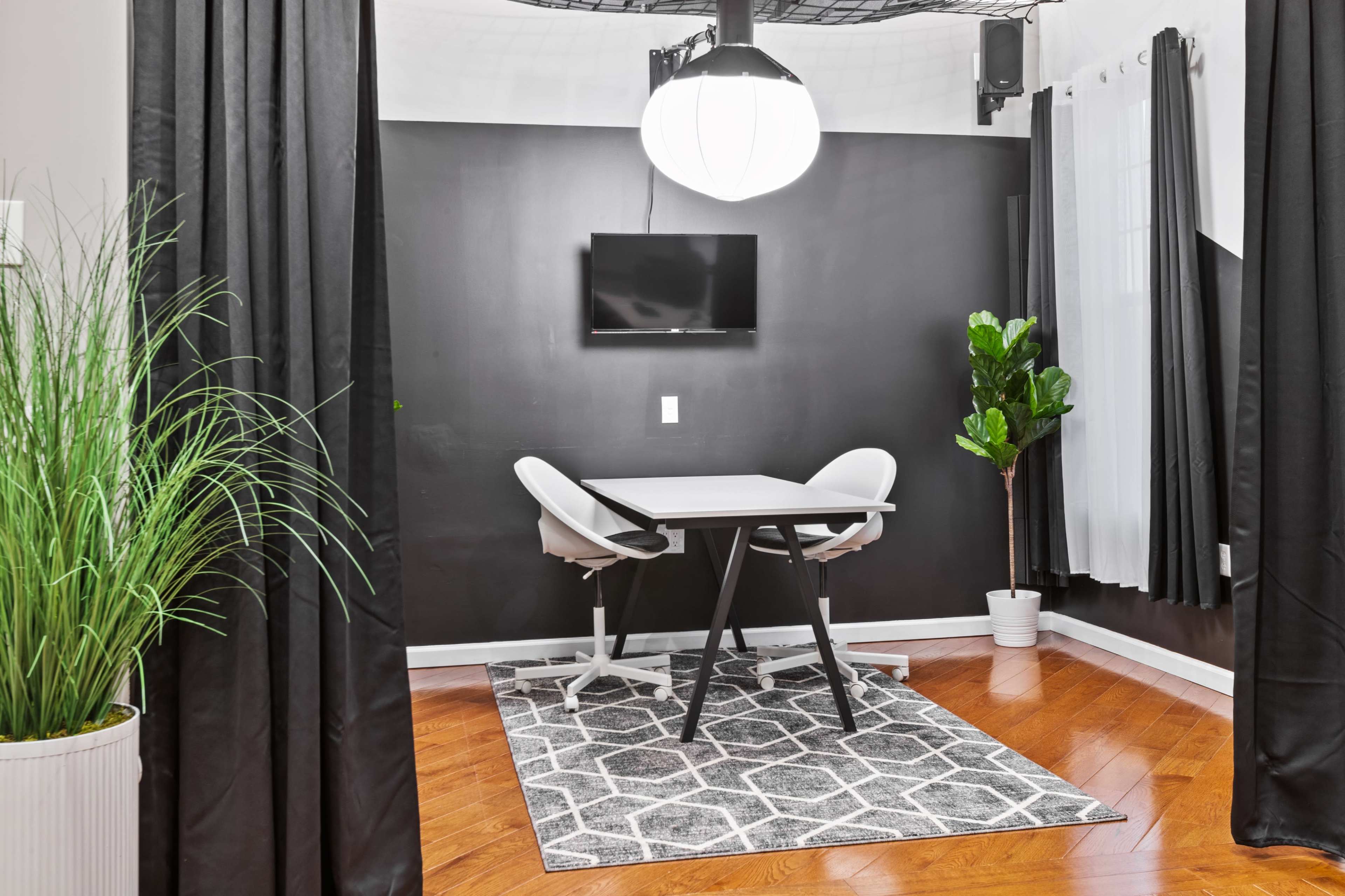 A small, modern workspace features a table with two chairs, a wall-mounted TV, and a potted plant, all enclosed by dark curtains.