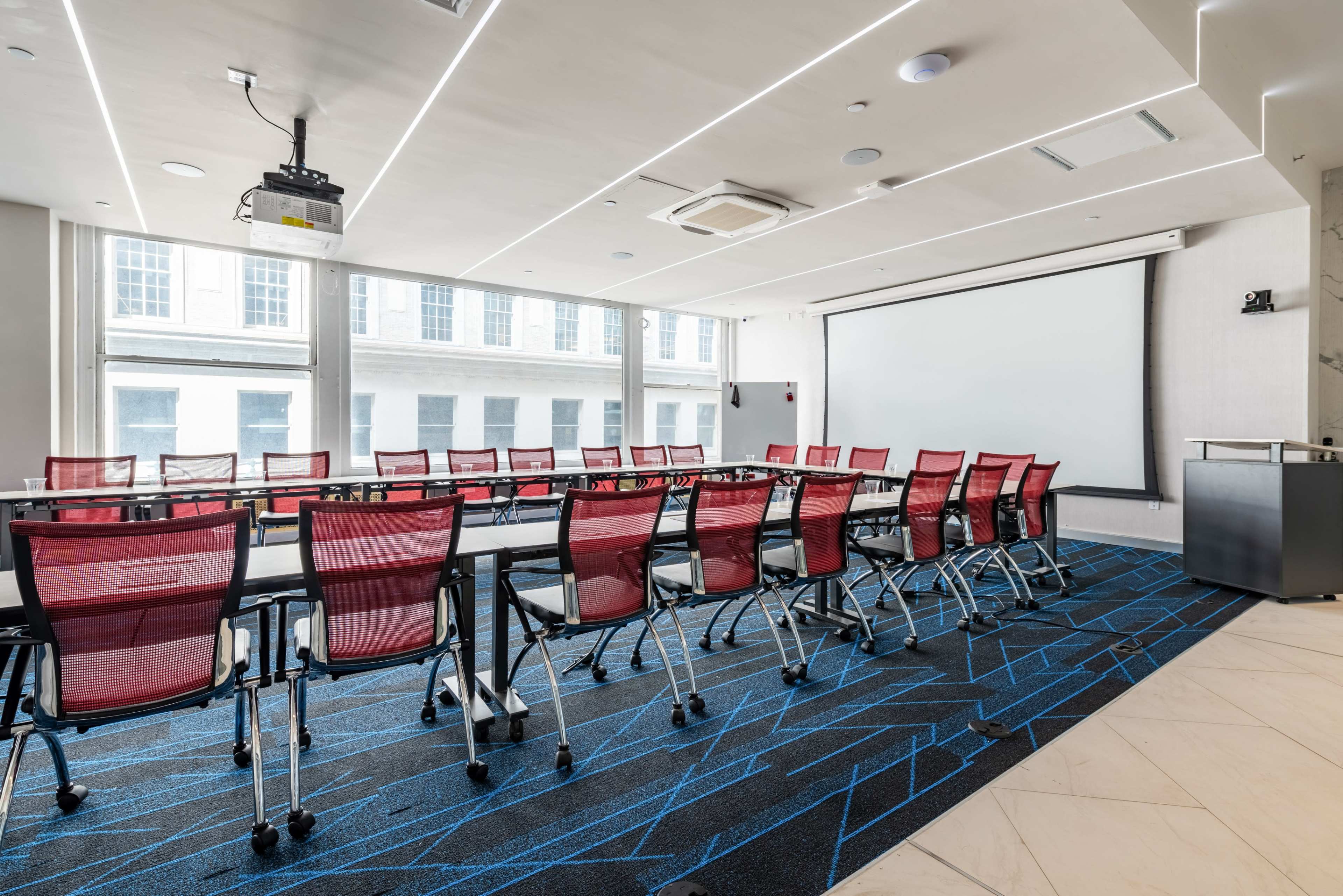 The image shows a modern conference room with a large rectangular table, red wheeled chairs, and a projection screen at one end.