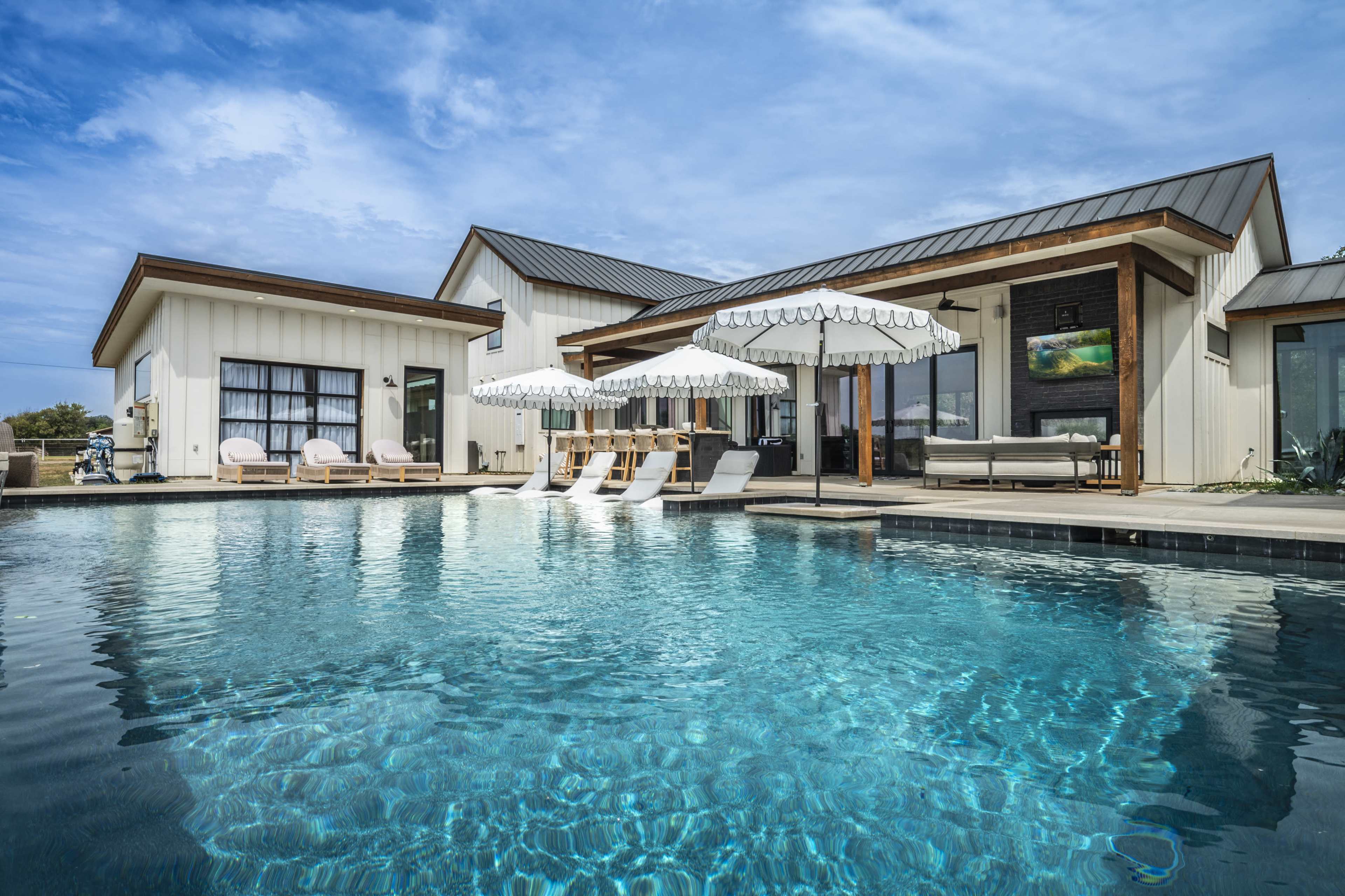 A modern house with a pool features several loungers and umbrellas beside the water, set against a clear blue sky.