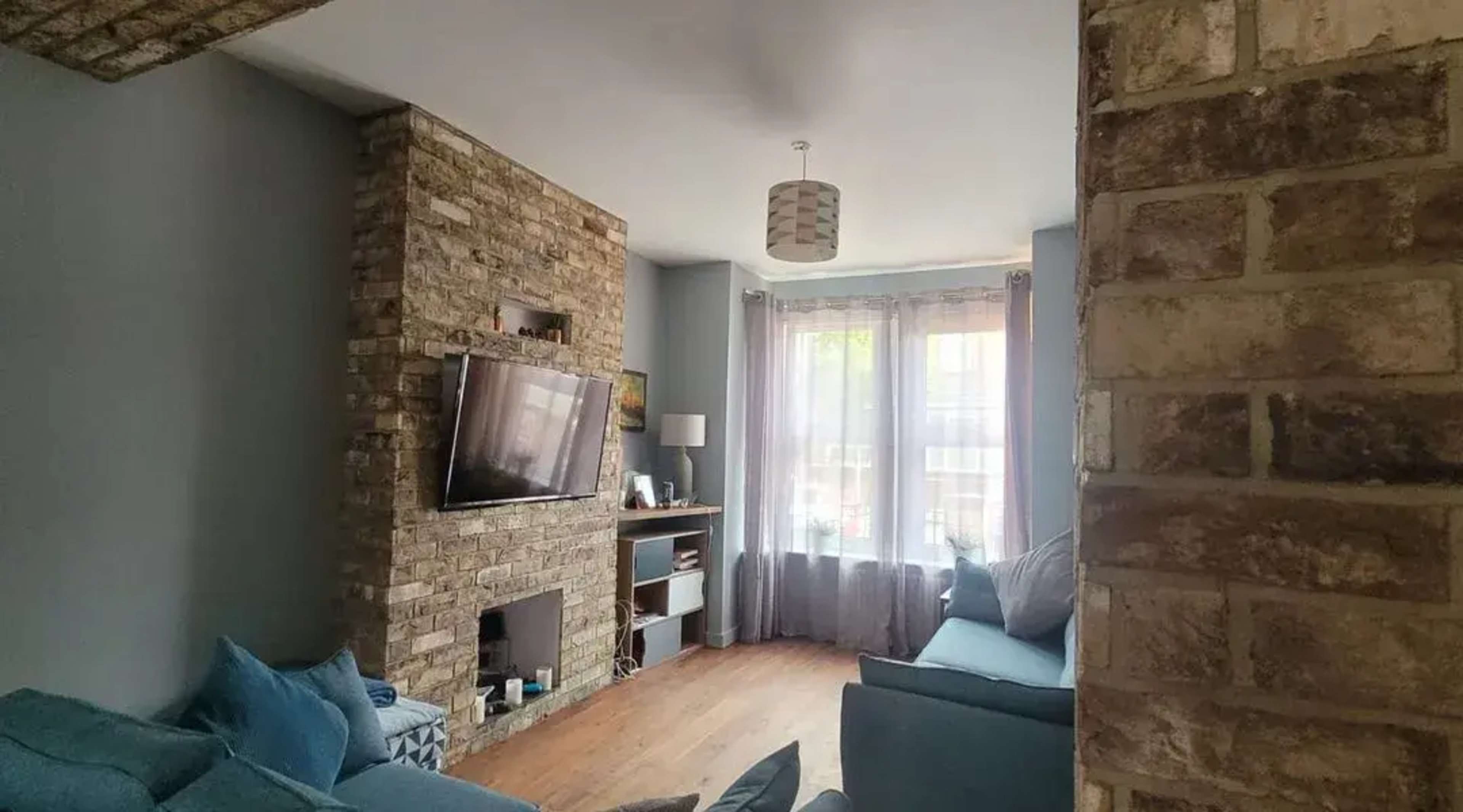 A cozy living room with a stone wall, a television mounted above a fireplace, and large windows draped with sheer curtains.