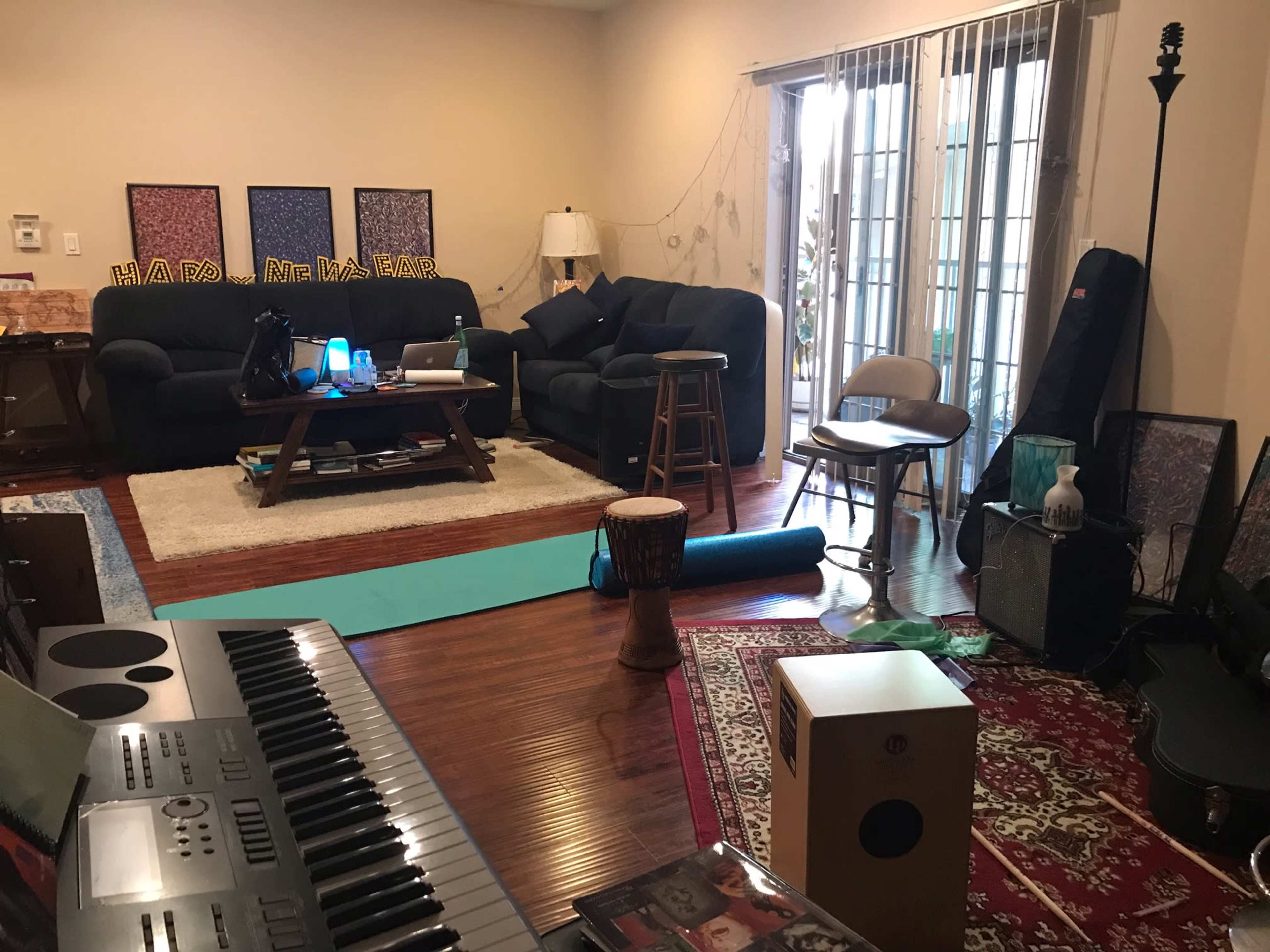 The living room features a keyboard, a coffee table with decorative items, a yoga mat, a drum, and seating arrangements near glass doors leading outside.