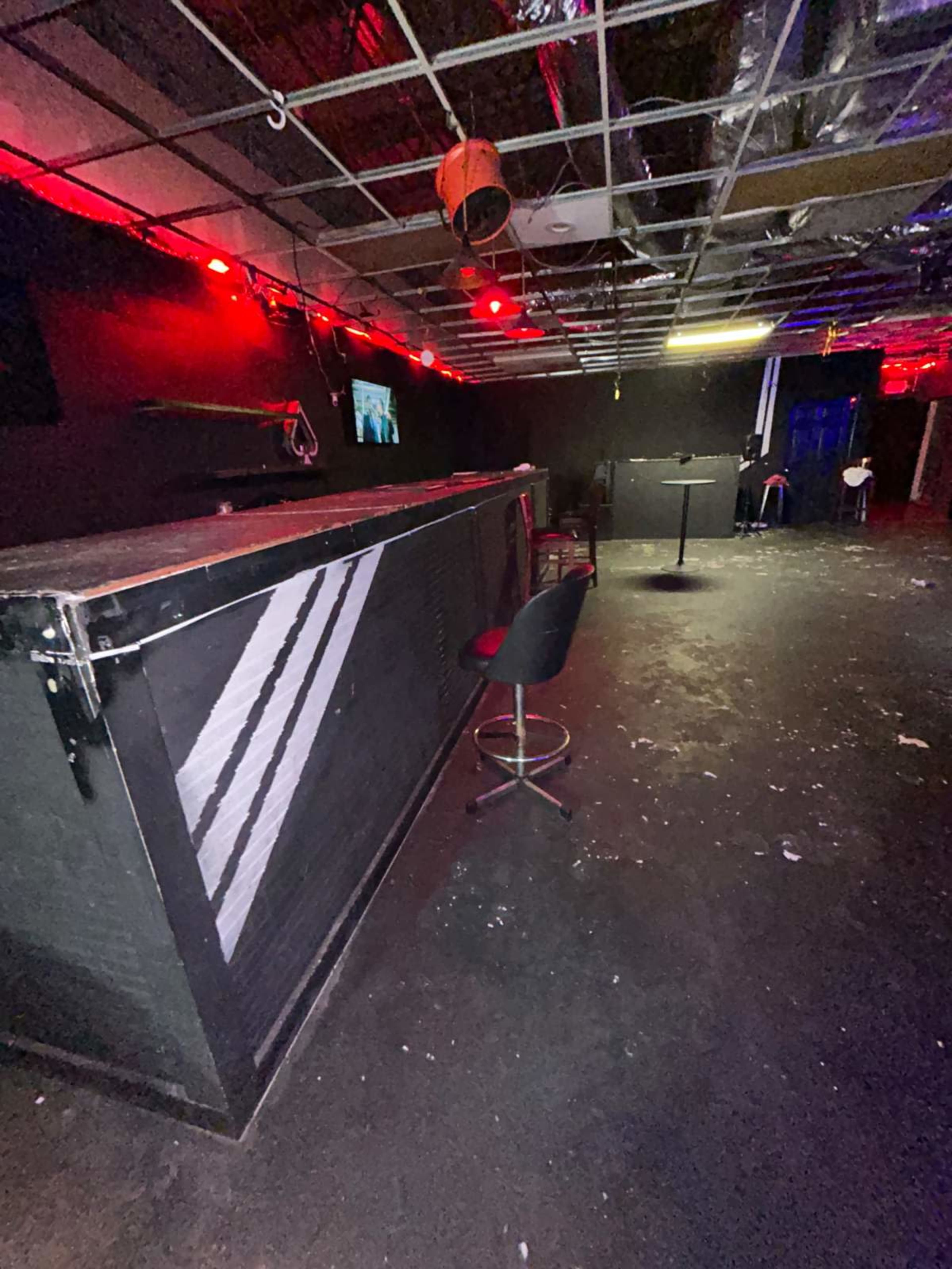 The image shows an empty bar area in a dimly lit setting, with red lighting and a partially ceiling-tiled ceiling.