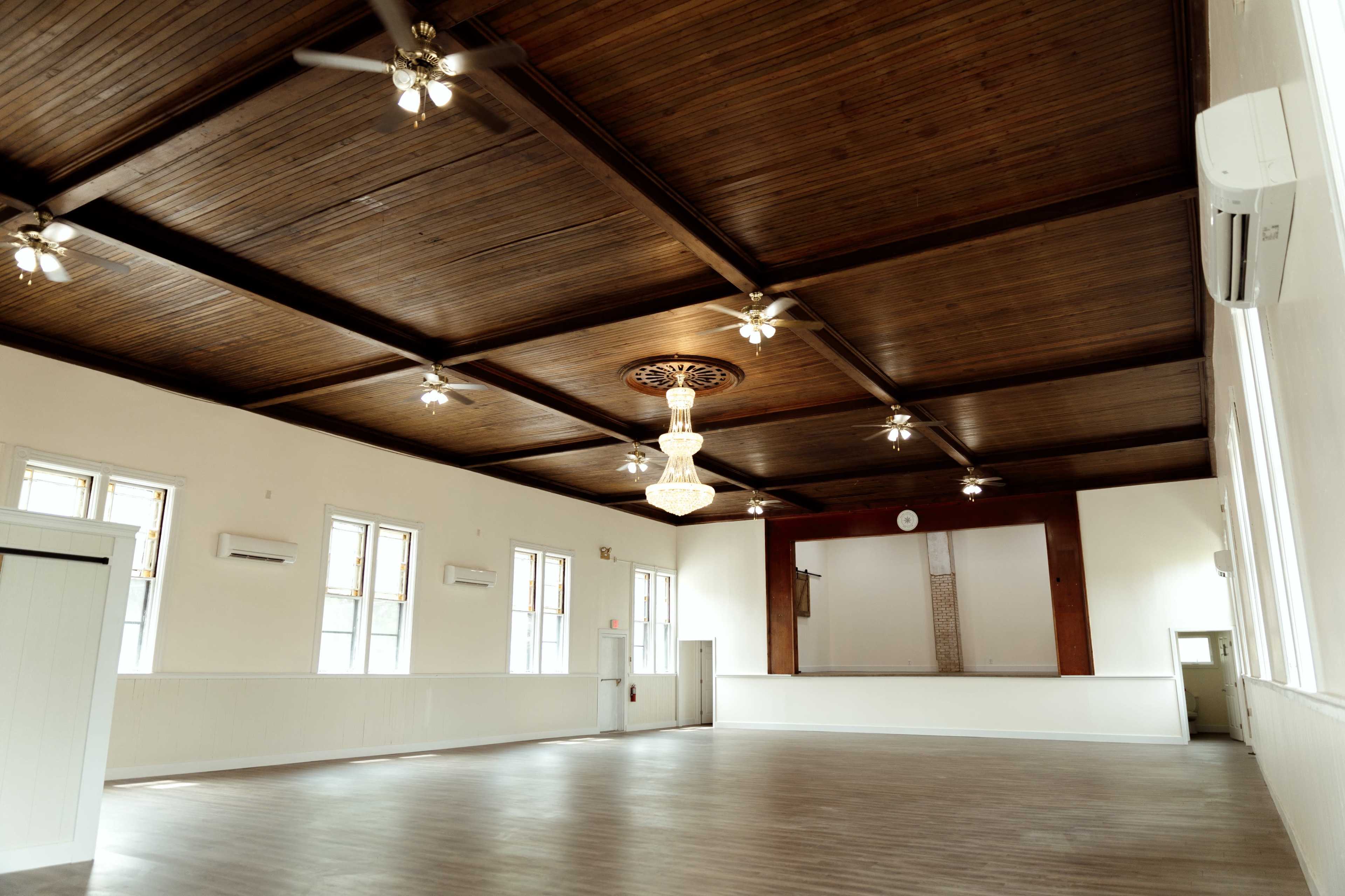 The image shows a spacious interior of a hall with wooden walls, a high ceiling with ceiling fans, and a central chandelier.