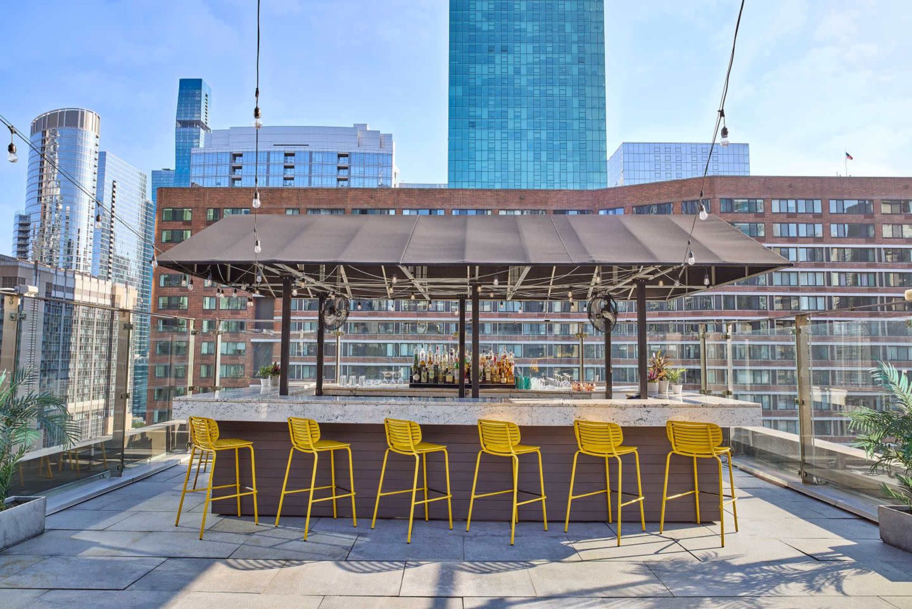 A rooftop bar features a large counter with yellow stools, surrounded by modern buildings and a clear blue sky.