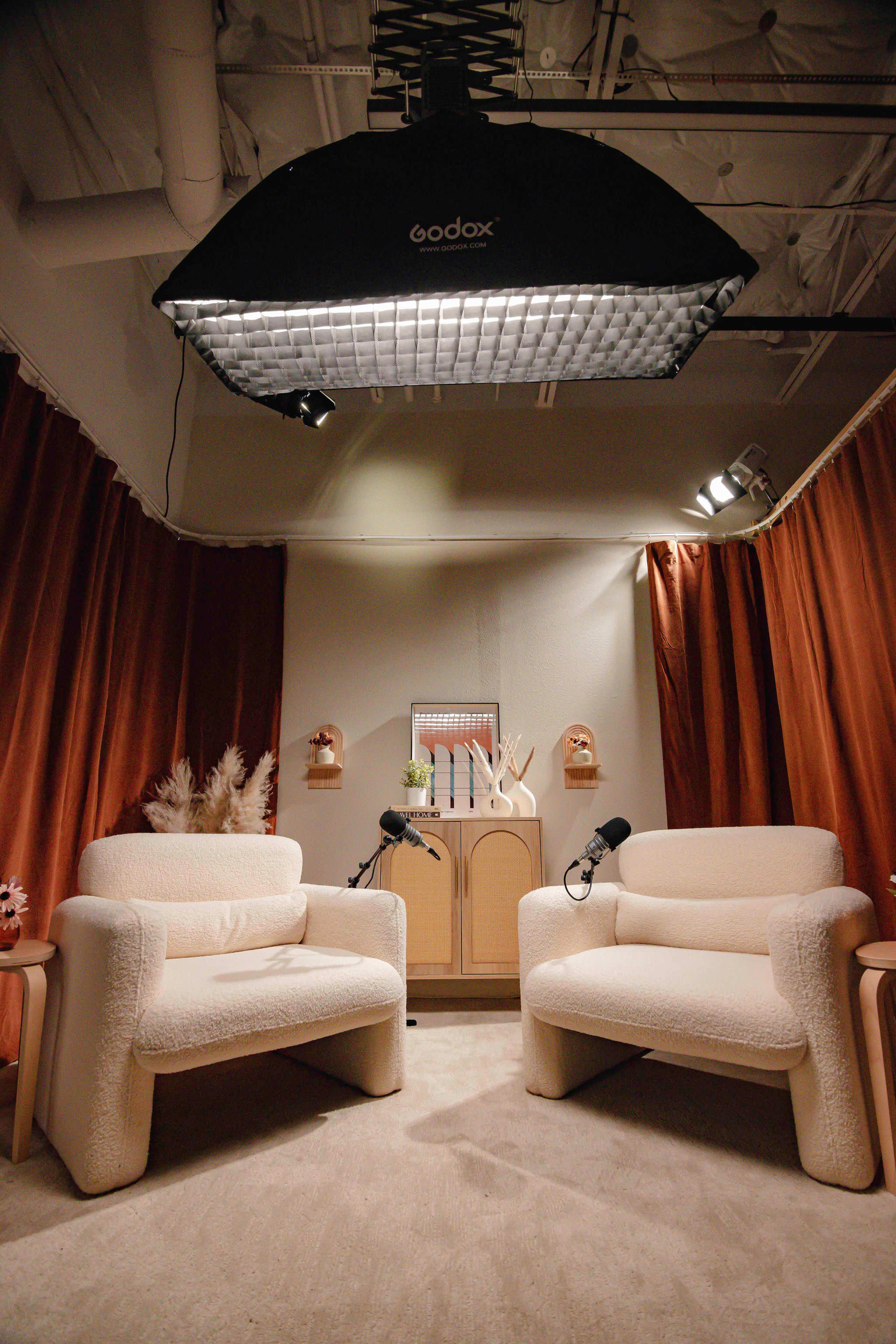 A cozy interview setup featuring two cream-colored armchairs, a wooden cabinet, and a large lighting softbox overhead, with microphones positioned on each chair.