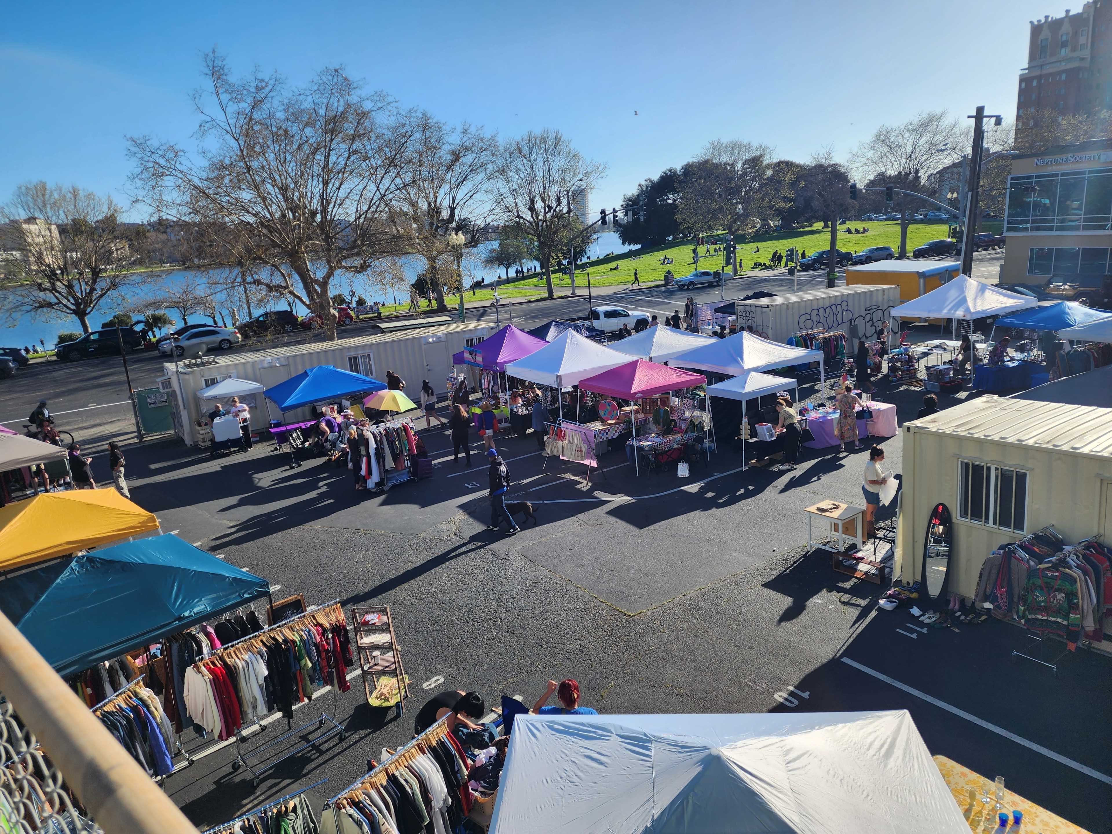A vibrant outdoor market features numerous vendors under colorful tents, with people browsing and shopping amid a backdrop of trees and a lake.