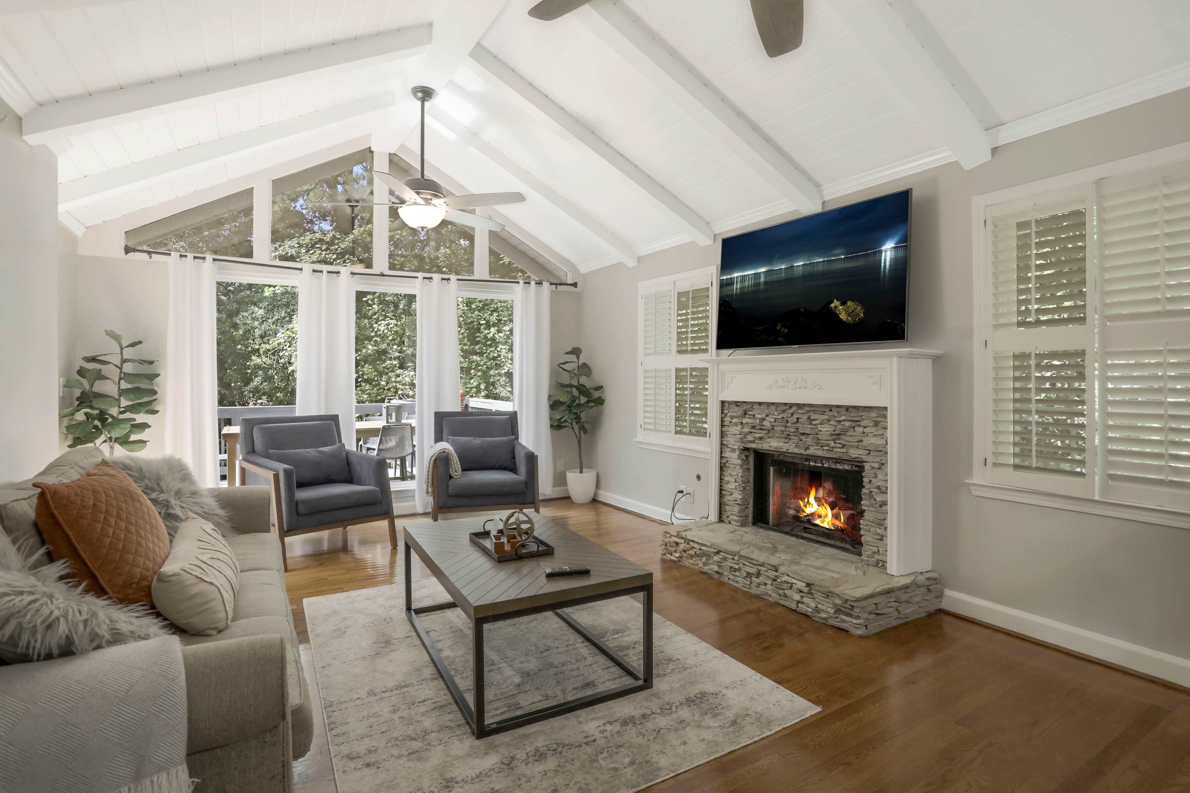A bright living room features a stone fireplace, a large television mounted above it, comfortable seating, and large windows overlooking a green view.