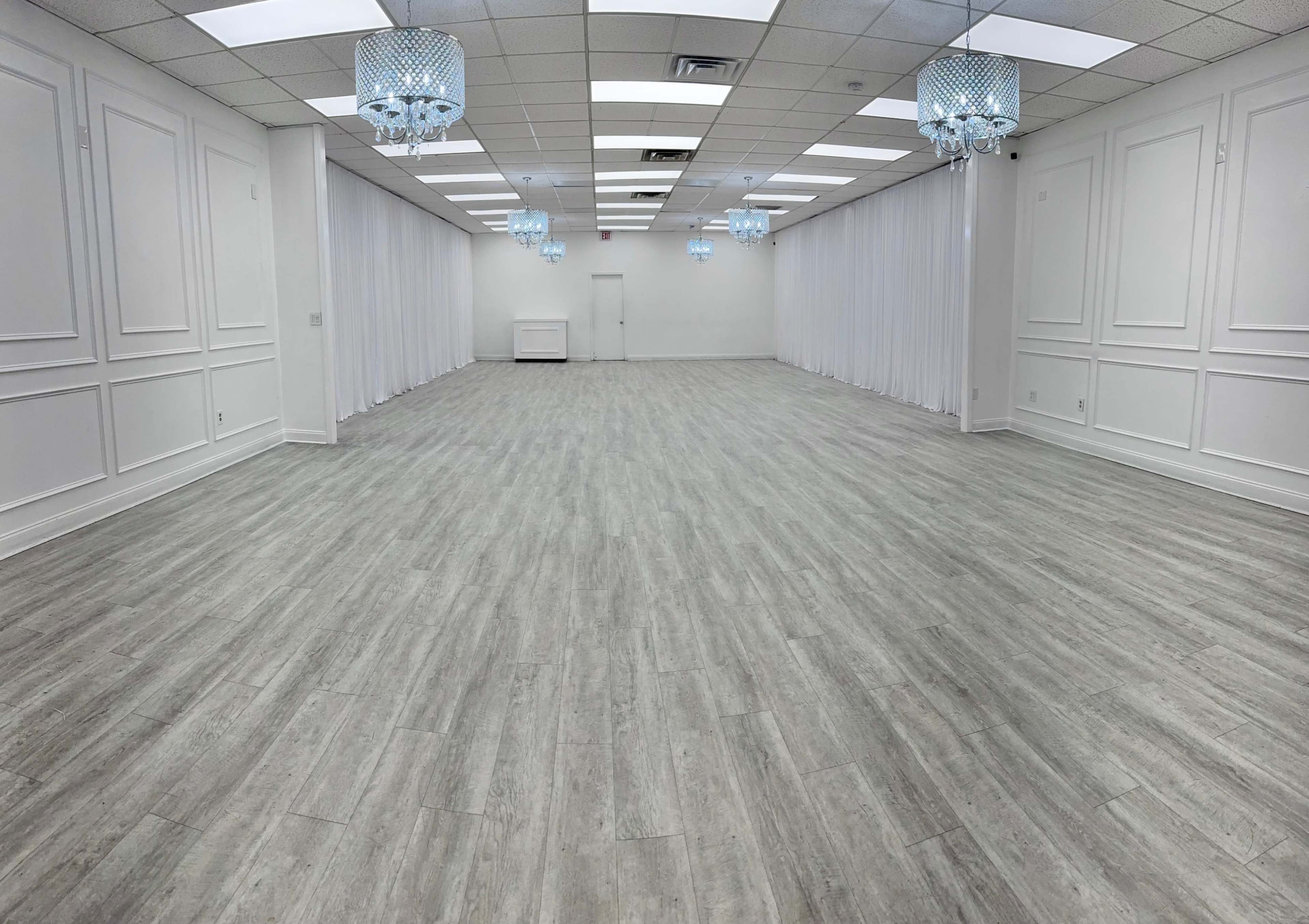 The image shows a spacious, empty room with light gray flooring, white walls, and decorative chandeliers hanging from the ceiling.