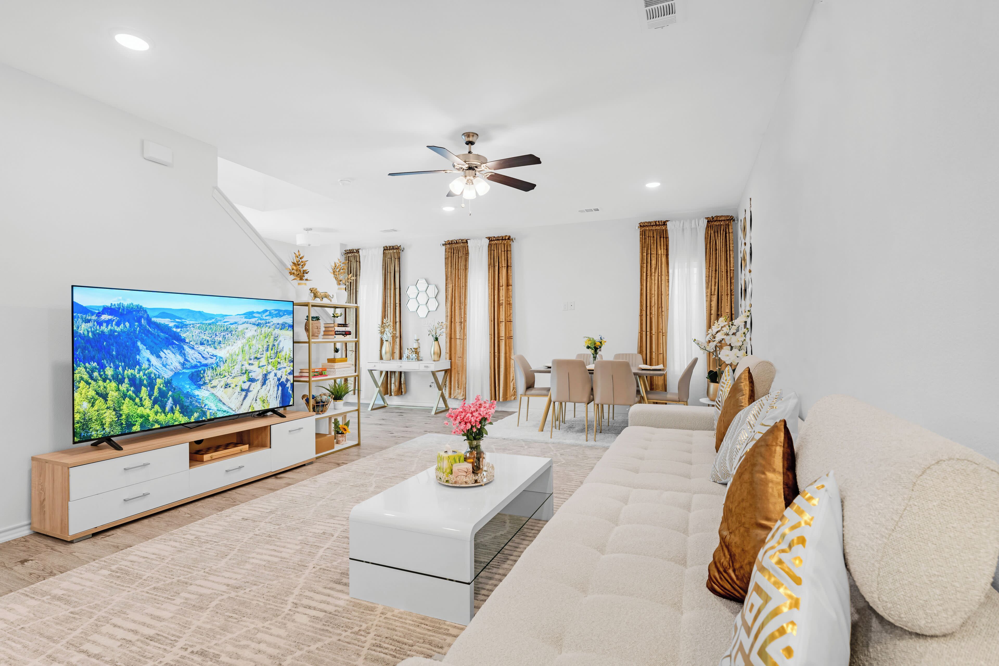 A modern living room with a large television, a beige sofa, a coffee table, and a dining area with floral arrangements and decorative accents.