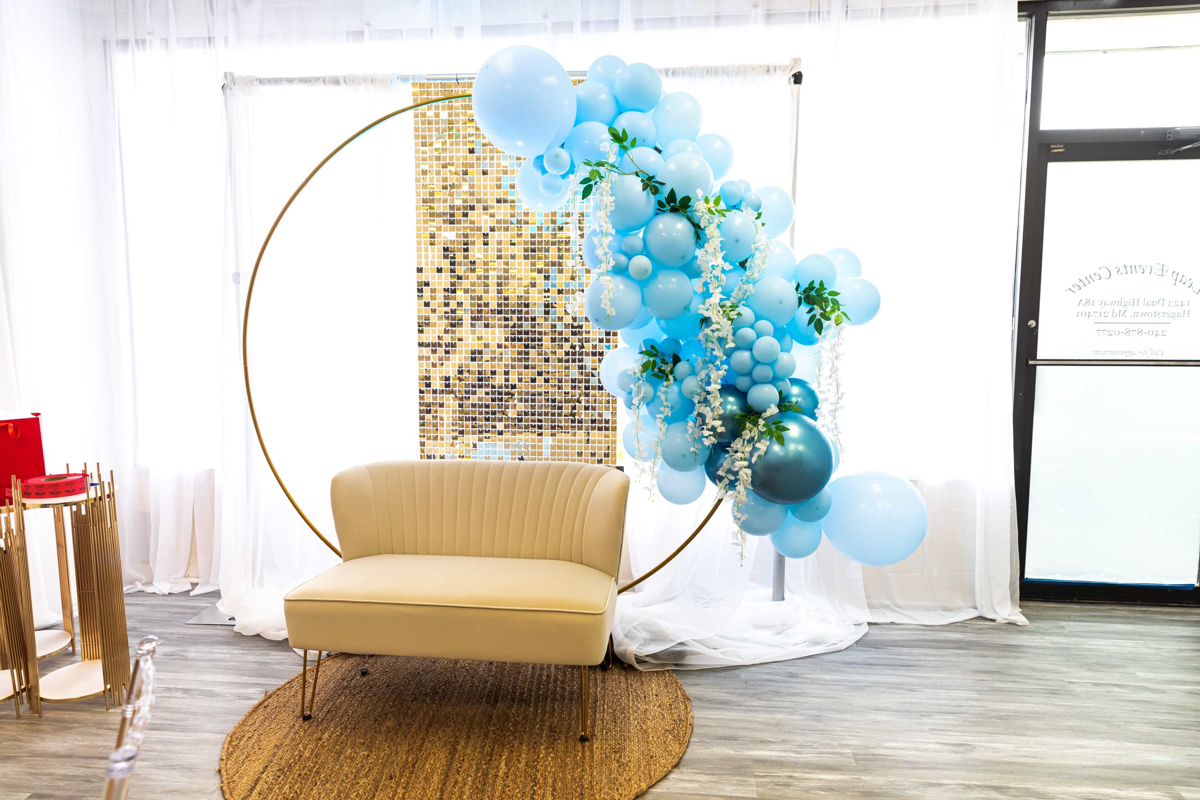 The scene features a light-colored sofa placed in front of a large circular balloon arrangement, with blue balloons and greenery against a backdrop of gold tiles.