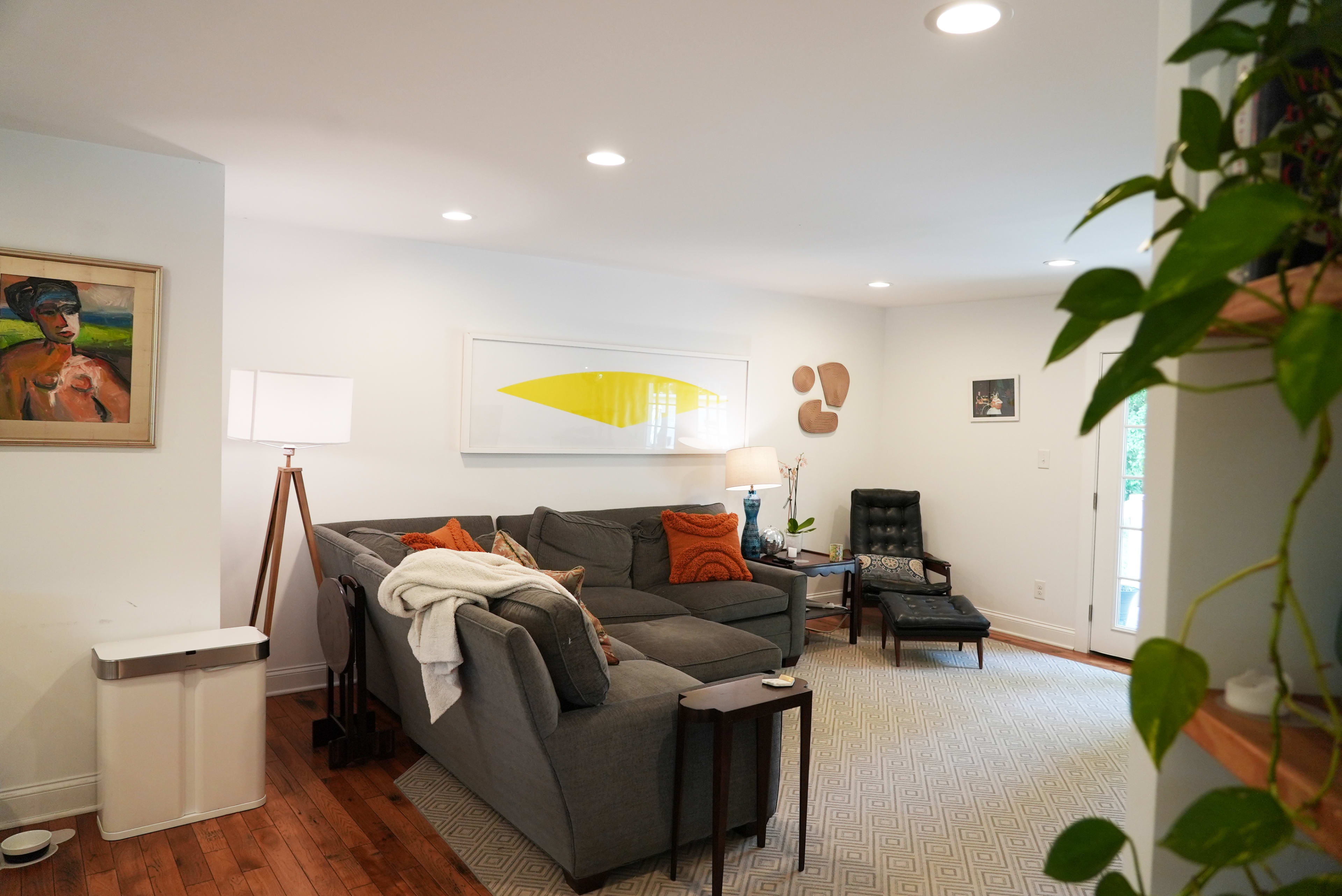 A modern living room features a gray sectional sofa with orange pillows, a large abstract wall art piece, and a black armchair, all illuminated by recessed lighting.