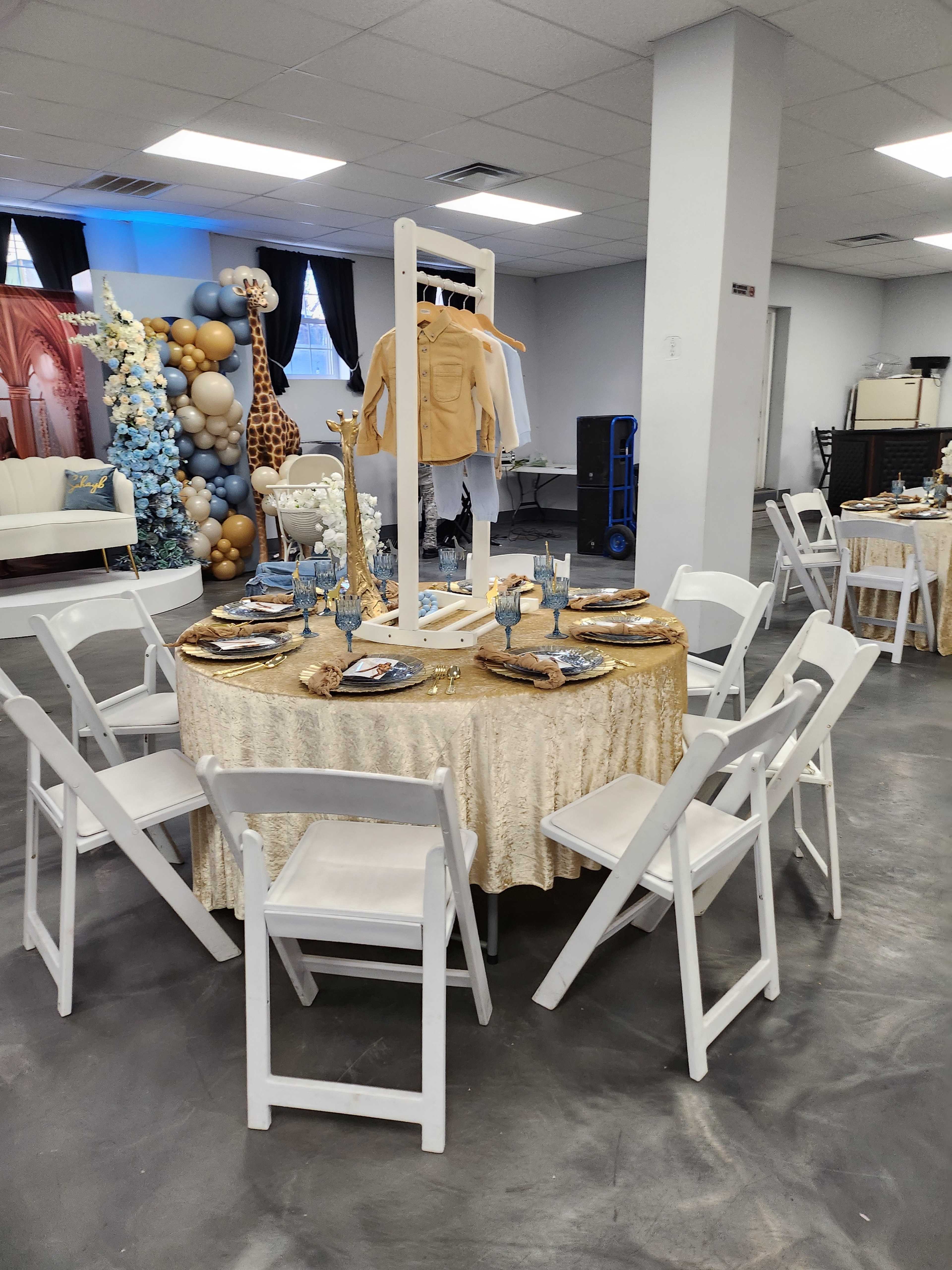 A round table covered with a gold tablecloth is set with plates and decorations, surrounded by white chairs in a banquet hall featuring a backdrop with balloons and decorative elements.