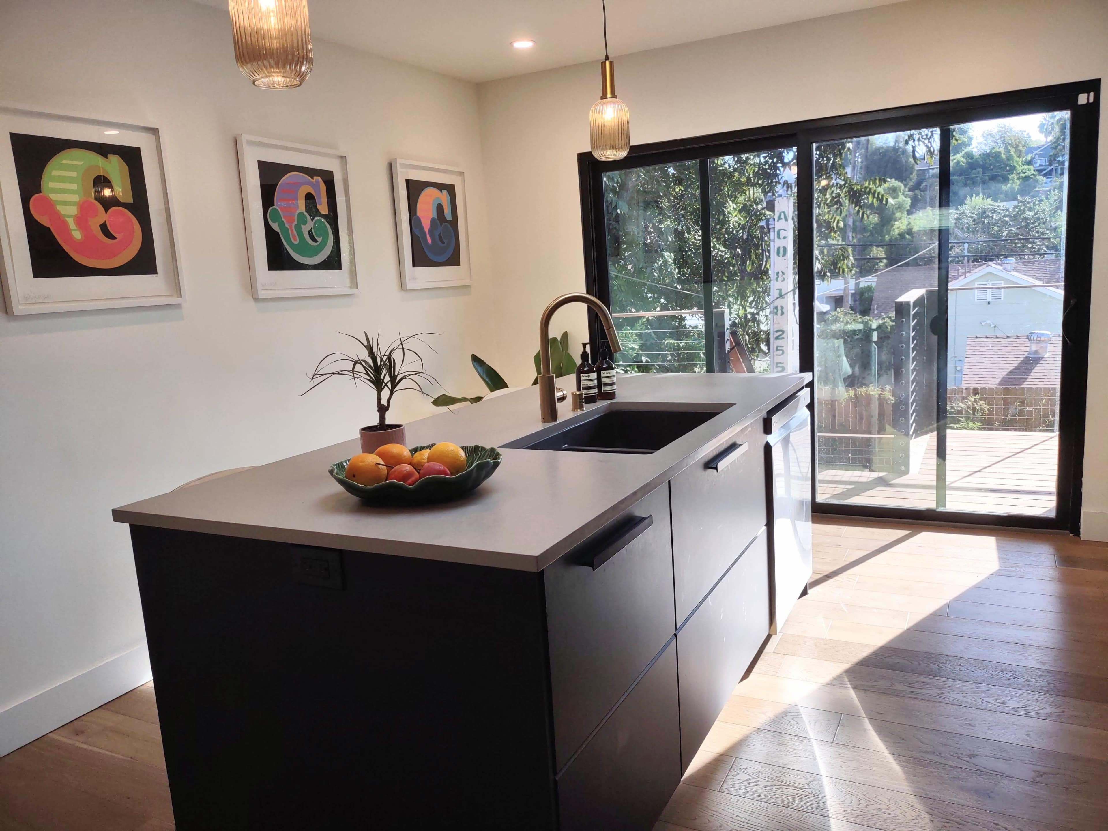 A modern kitchen features an island with a sink and a bowl of fruit, complemented by framed art on the walls and a view of a deck through sliding glass doors.