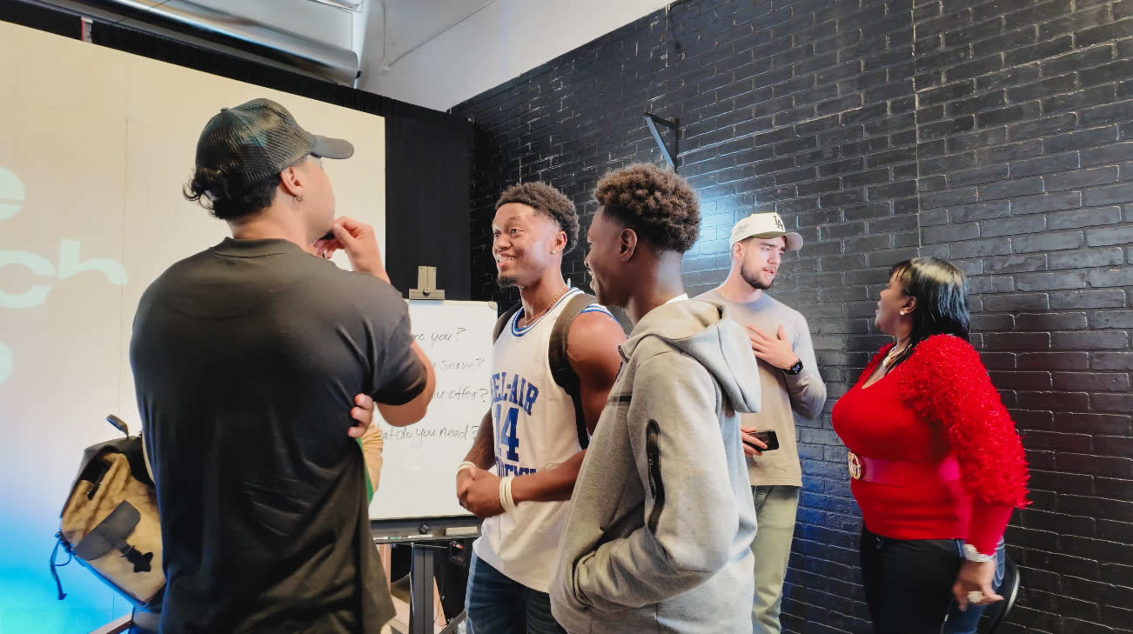 A group of young adults engages in conversation in a room with black brick walls and a whiteboard.