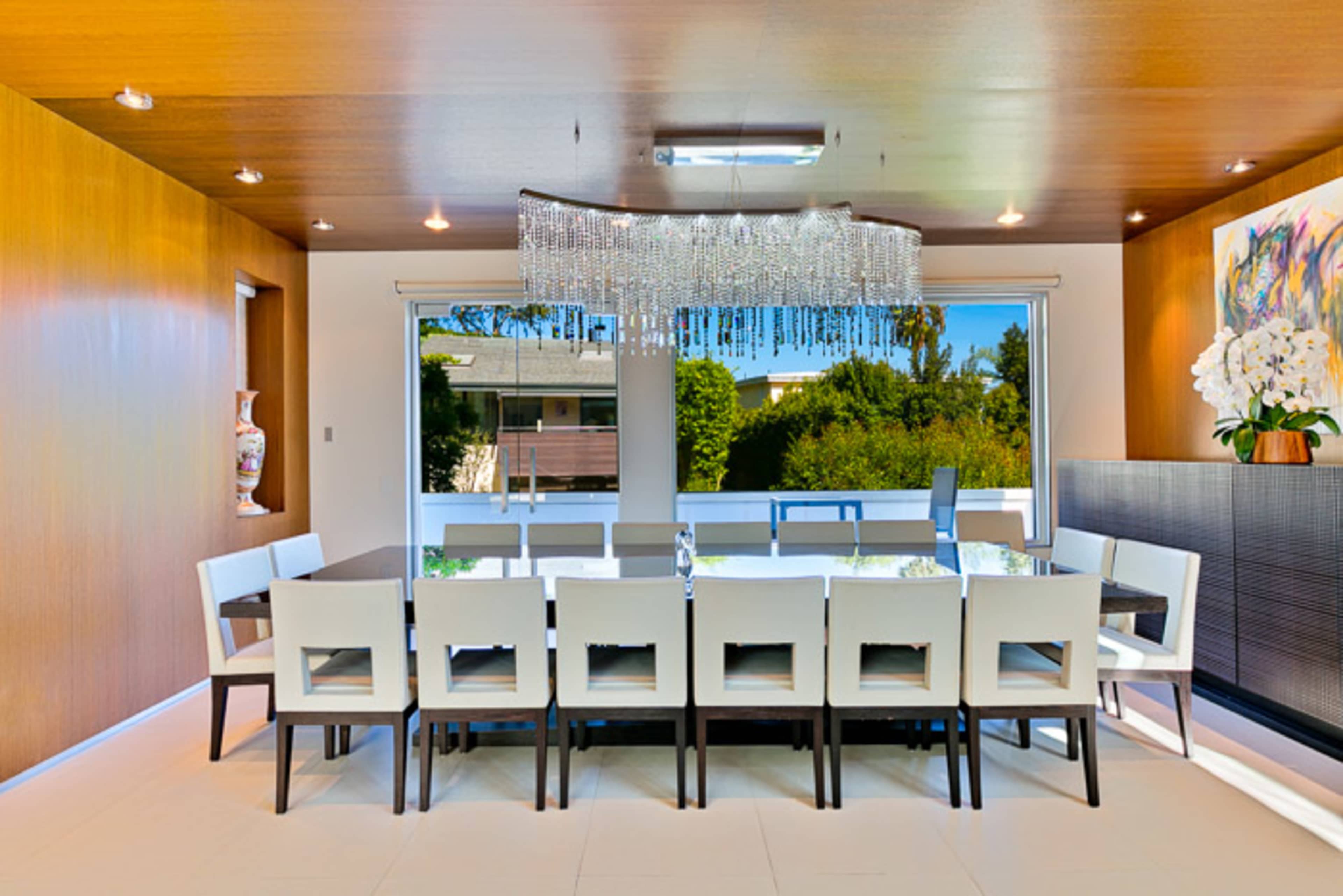 The image shows a modern dining room with a large rectangular table surrounded by white chairs, illuminated by a crystal chandelier and featuring large windows that overlook greenery outside.