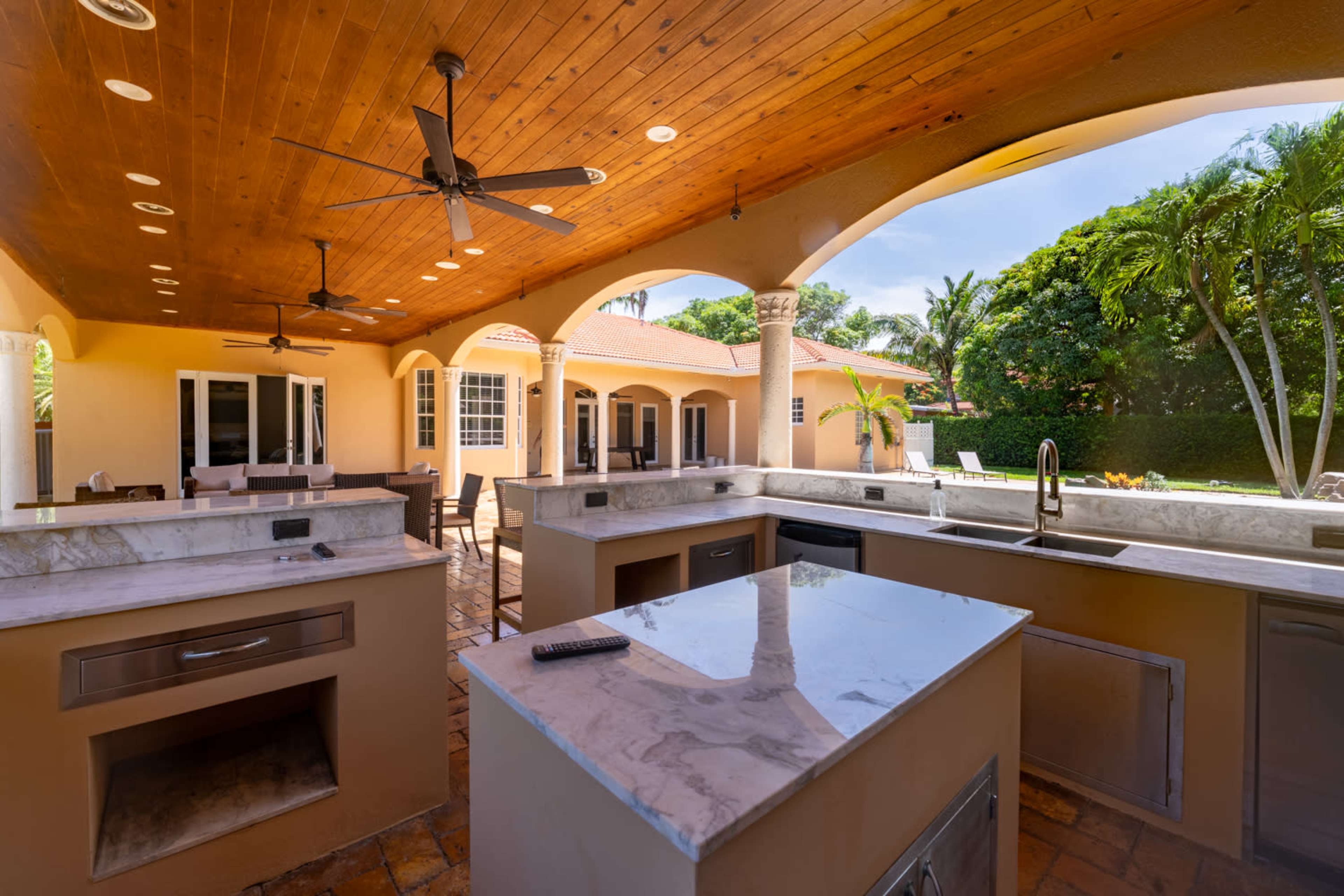 The image shows an outdoor kitchen featuring a marble countertop, built-in grills, and a spacious area with a wooden ceiling and ceiling fans, surrounded by tropical landscaping.