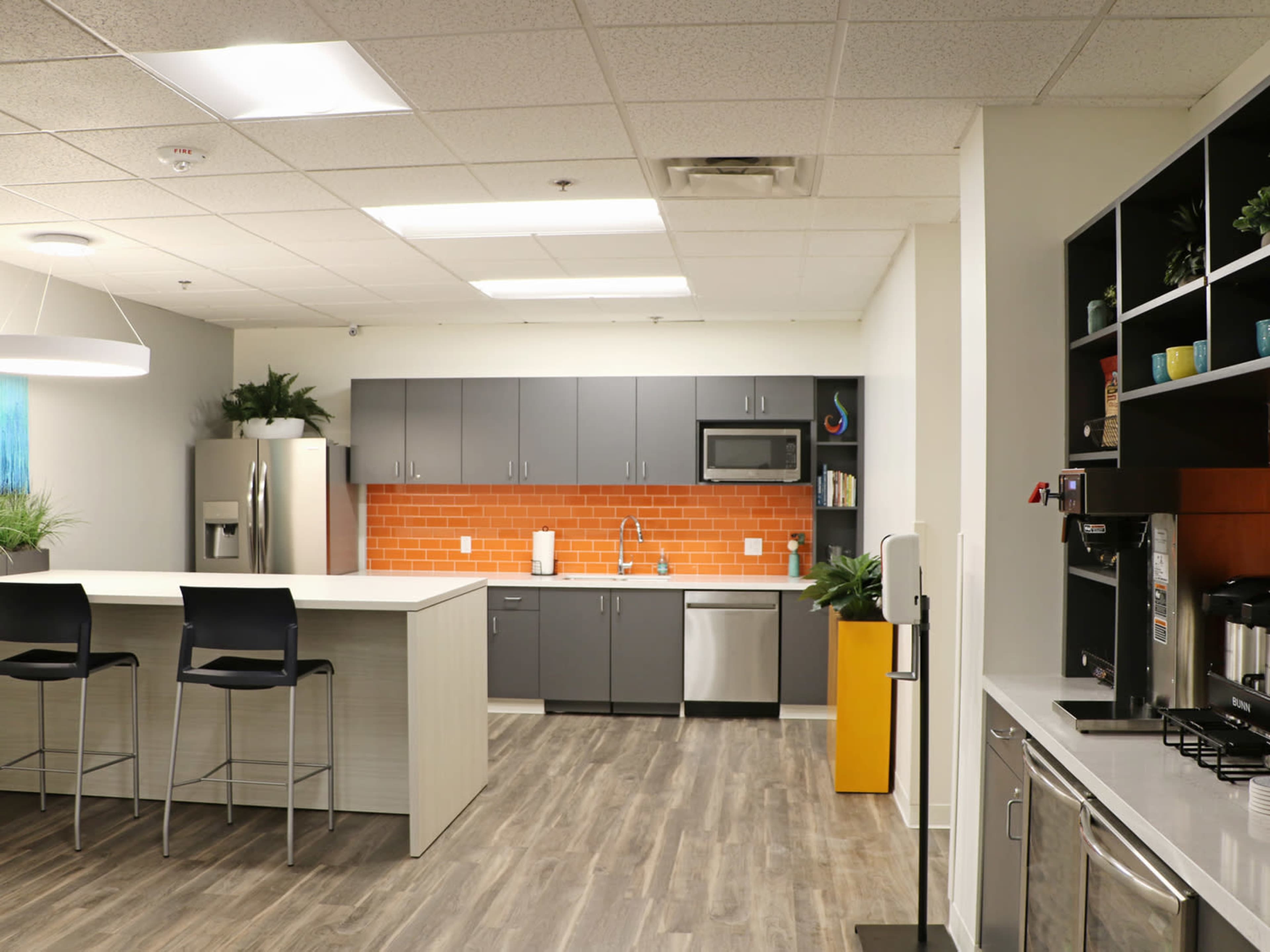 A modern kitchen features gray cabinetry, stainless steel appliances, and a central island with bar stools, accented by an orange tiled backsplash.